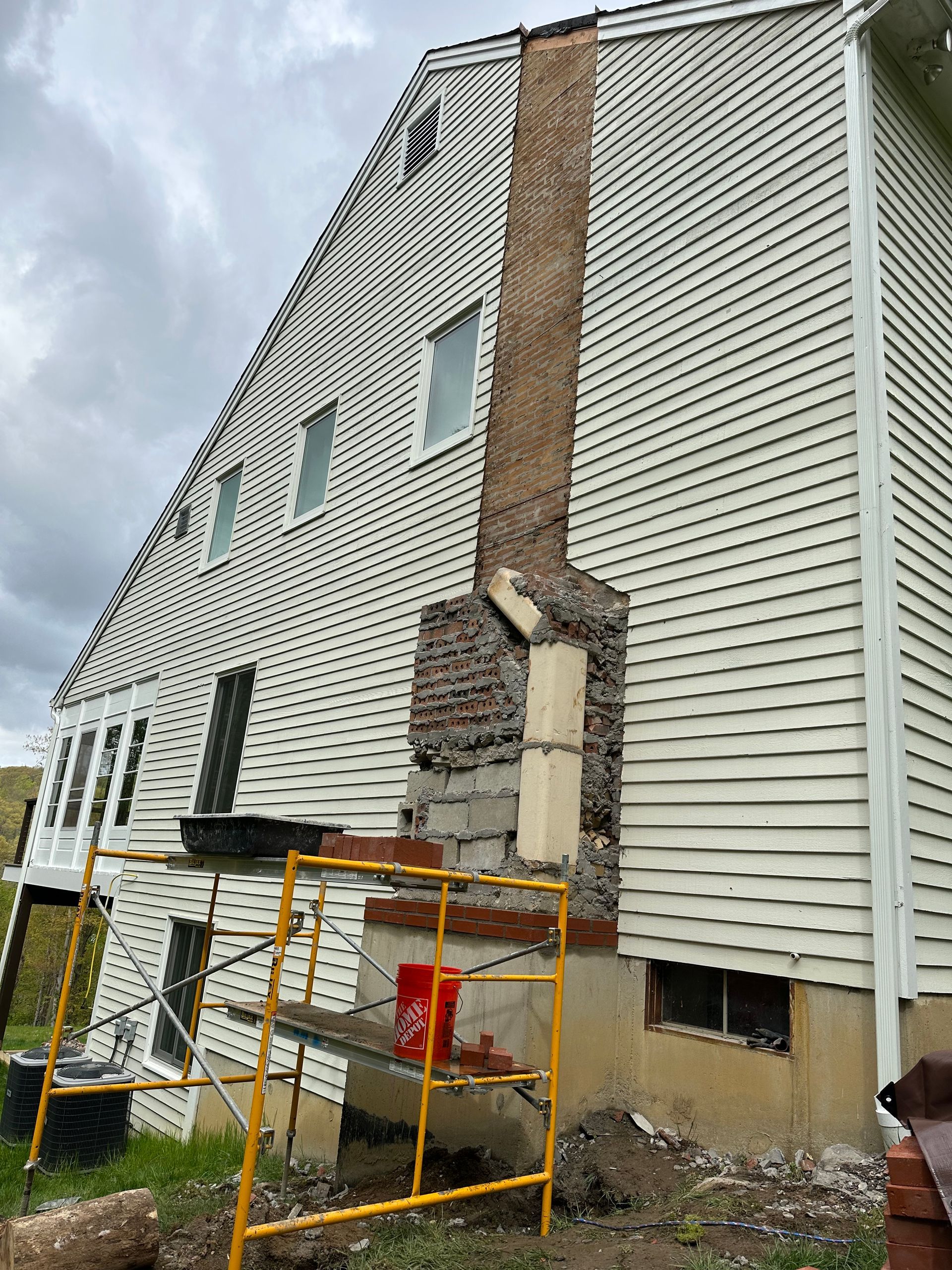 A side view of a house undergoing masonry repairs on a tall chimney, with yellow scaffolding set up at the base.