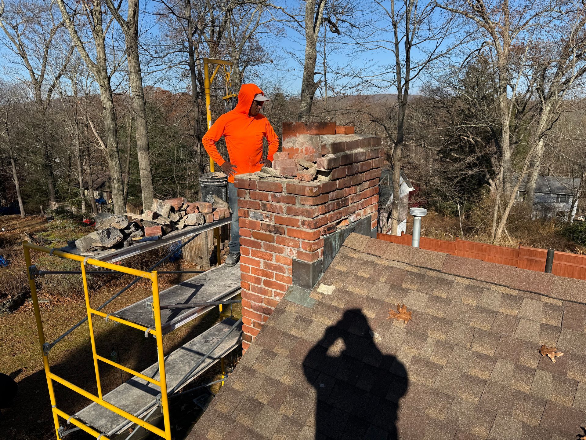 A person in an orange hoodie stands on scaffolding next to a brick chimney under repair on a shingled roof.