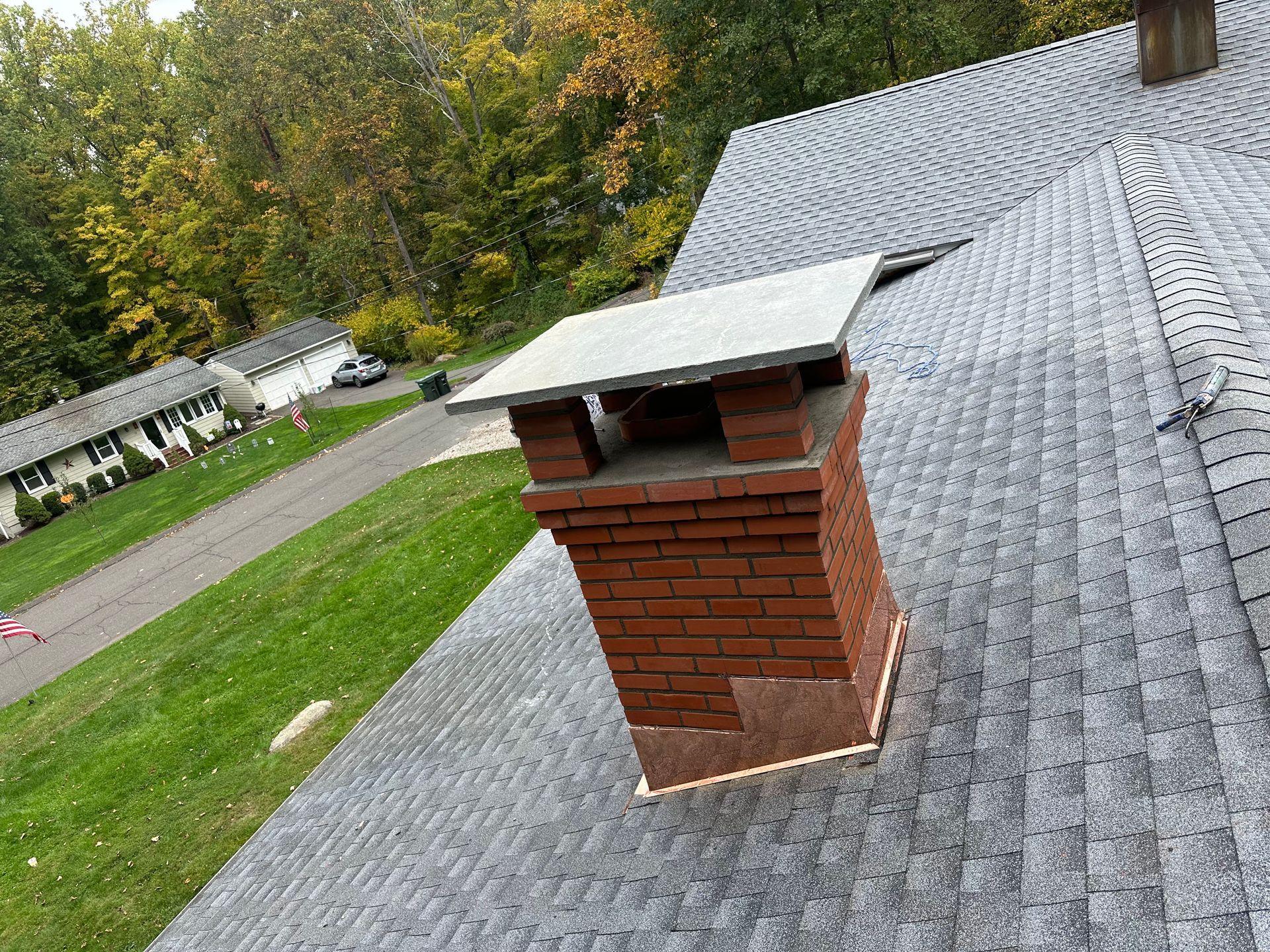 A brick chimney with a metal cap centered on a gray shingled roof, overlooking a suburban street and green trees.