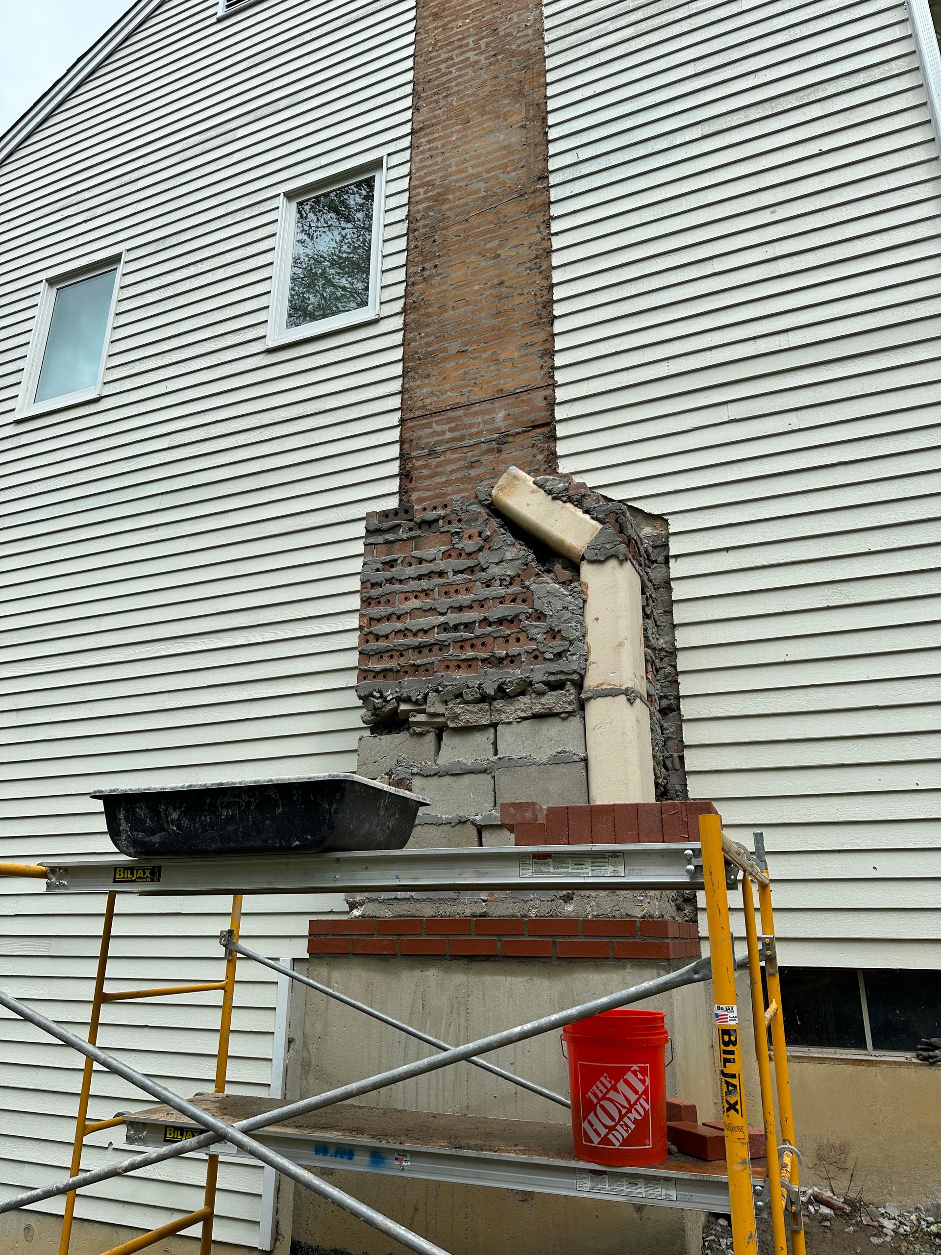 A chimney undergoing repairs with exposed brick and concrete blocks, partially covered by scaffolding and a red bucket.