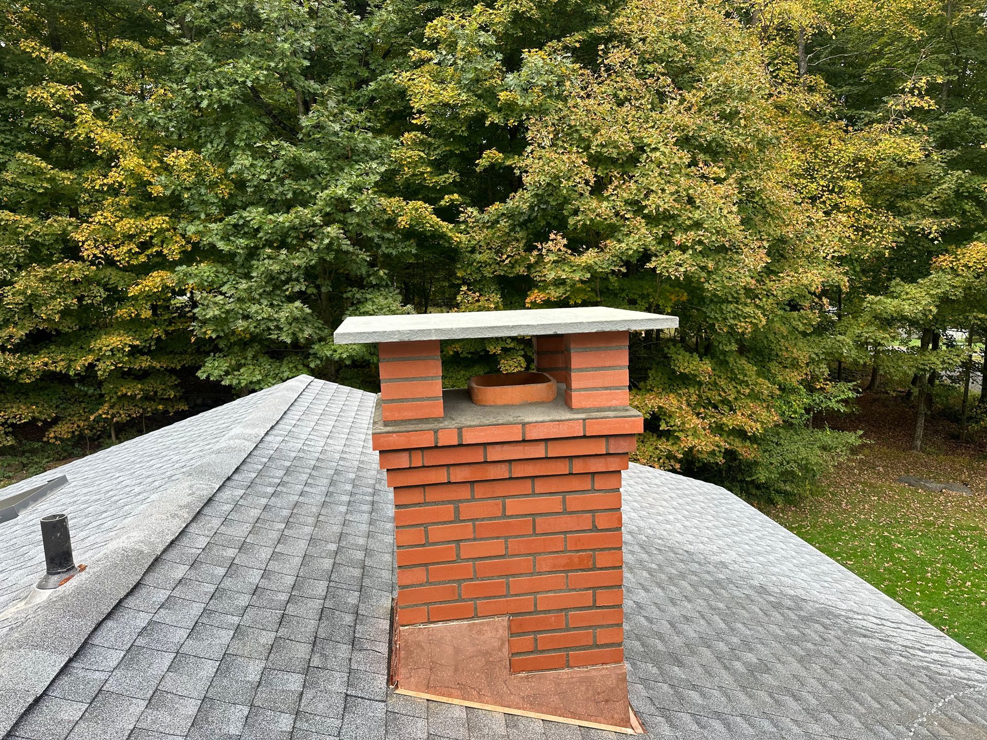 A red brick chimney with a concrete cap sits on a grey shingled roof against a background of autumn trees.