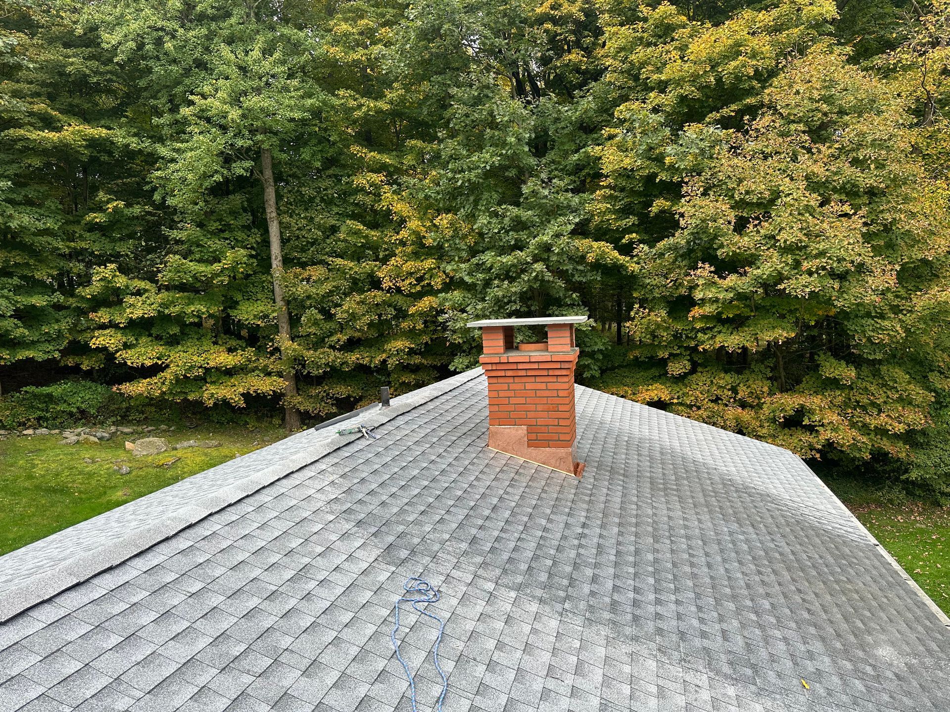A red brick chimney standing on a grey shingled roof, set against a backdrop of trees with early autumn foliage.