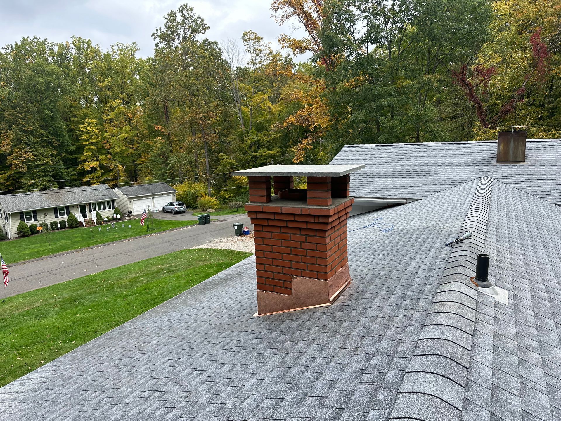 A brick chimney with a concrete cap sits on a grey shingled roof, overlooking a suburban street and trees.