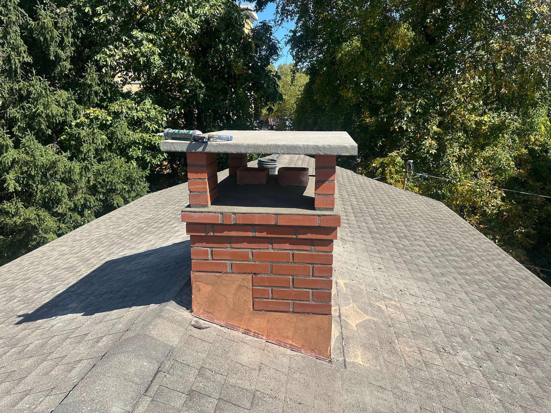 A brick chimney with a concrete cap stands on a shingled residential roof, surrounded by green trees on a sunny day.