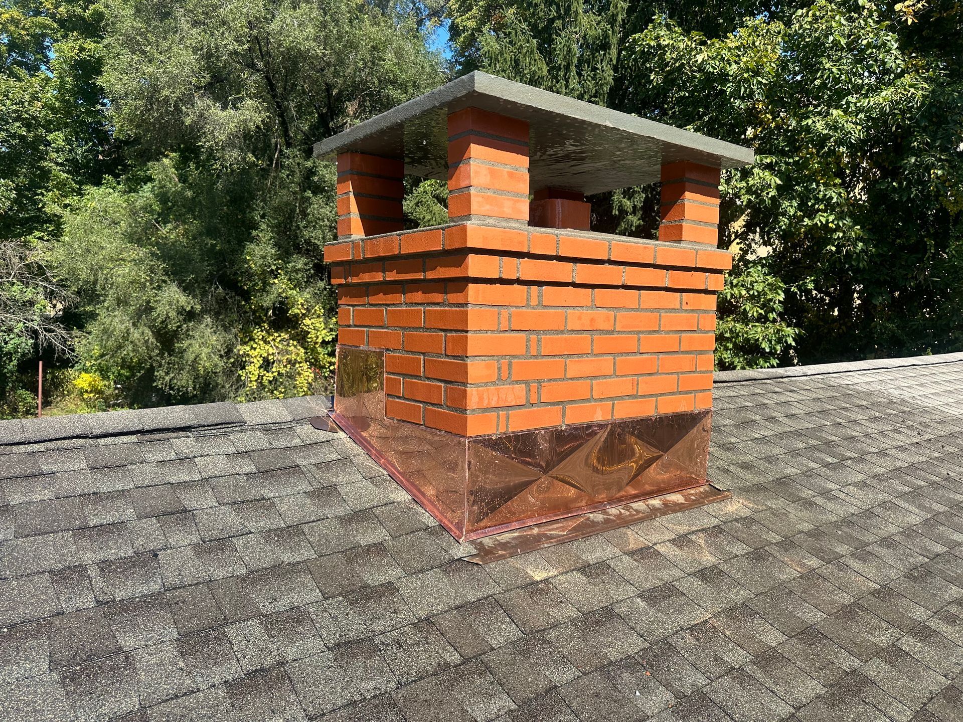 A brick chimney on a shingled roof with copper flashing and a protective cap, surrounded by trees.