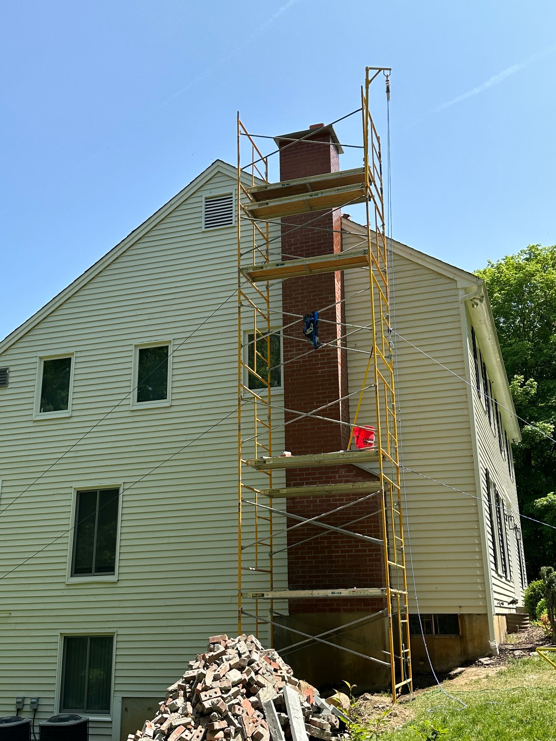 Tall scaffolding stands against a house, reaching a brick chimney under repair, with a pile of bricks on the ground below.