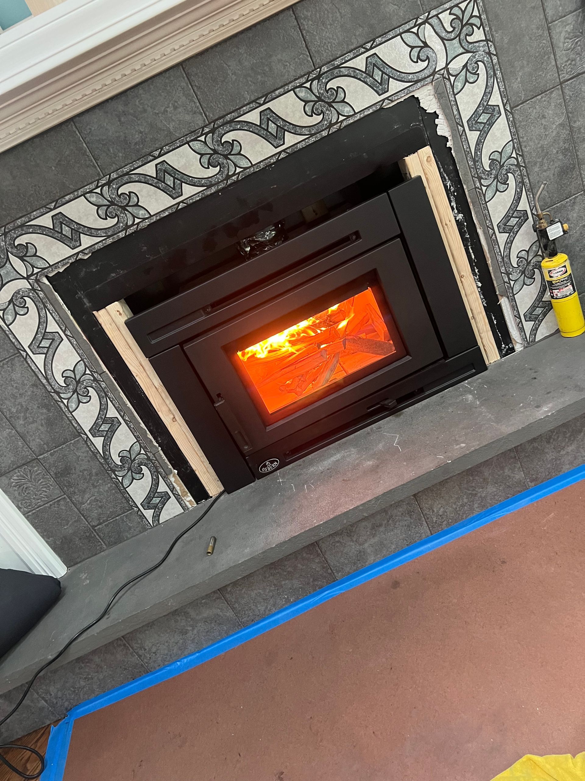A wood-burning stove insert with a bright fire, framed by decorative gray tiles and unfinished wood trim.