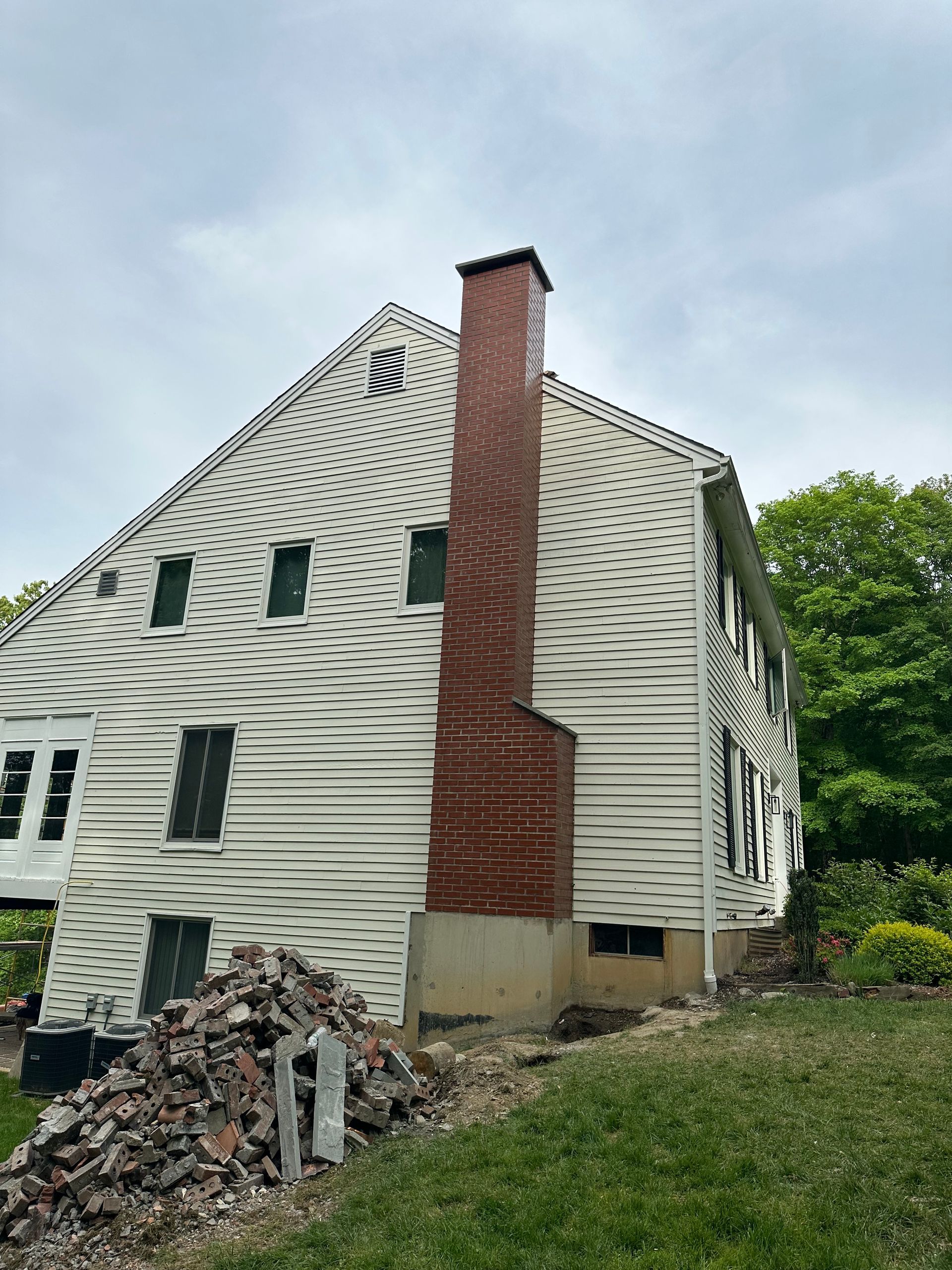 A side view of a house with warped, melted light-colored siding, a large brick chimney, and a pile of debris on the ground.