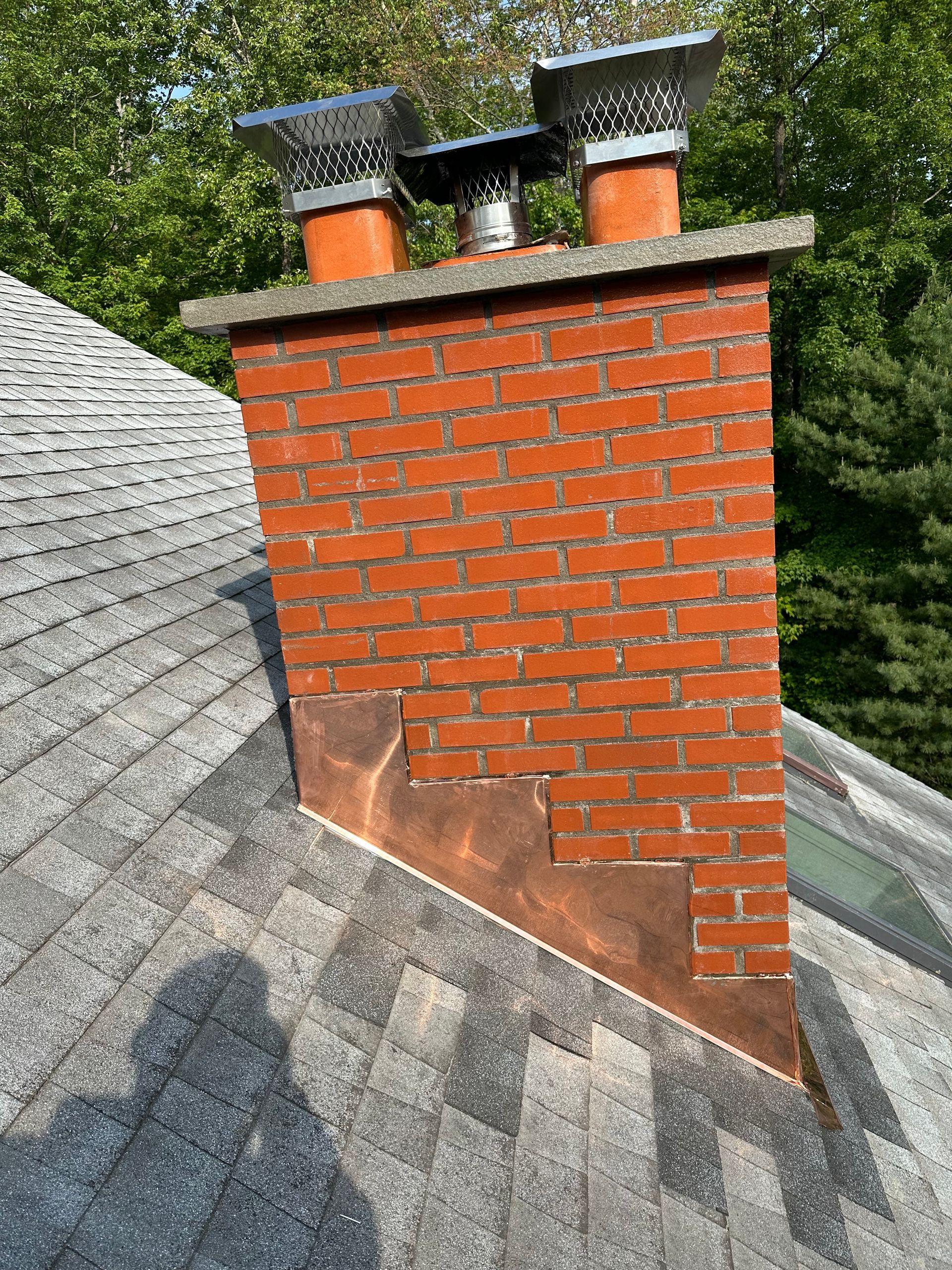 A red brick chimney with three metal flue caps and copper step flashing on a gray shingled roof.