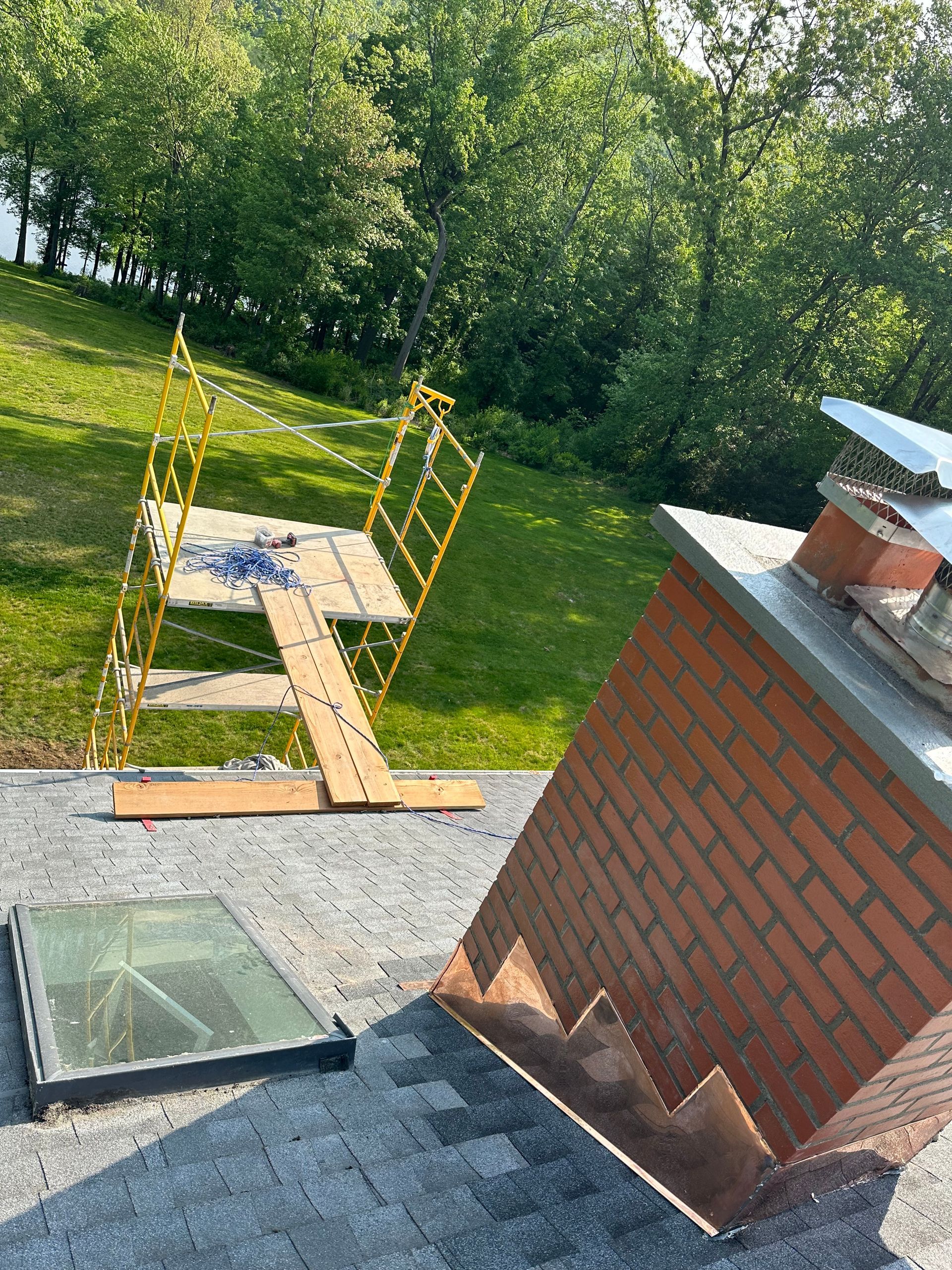 A red brick chimney with metal flashing on a grey shingled roof next to a skylight, with yellow scaffolding in the yard.