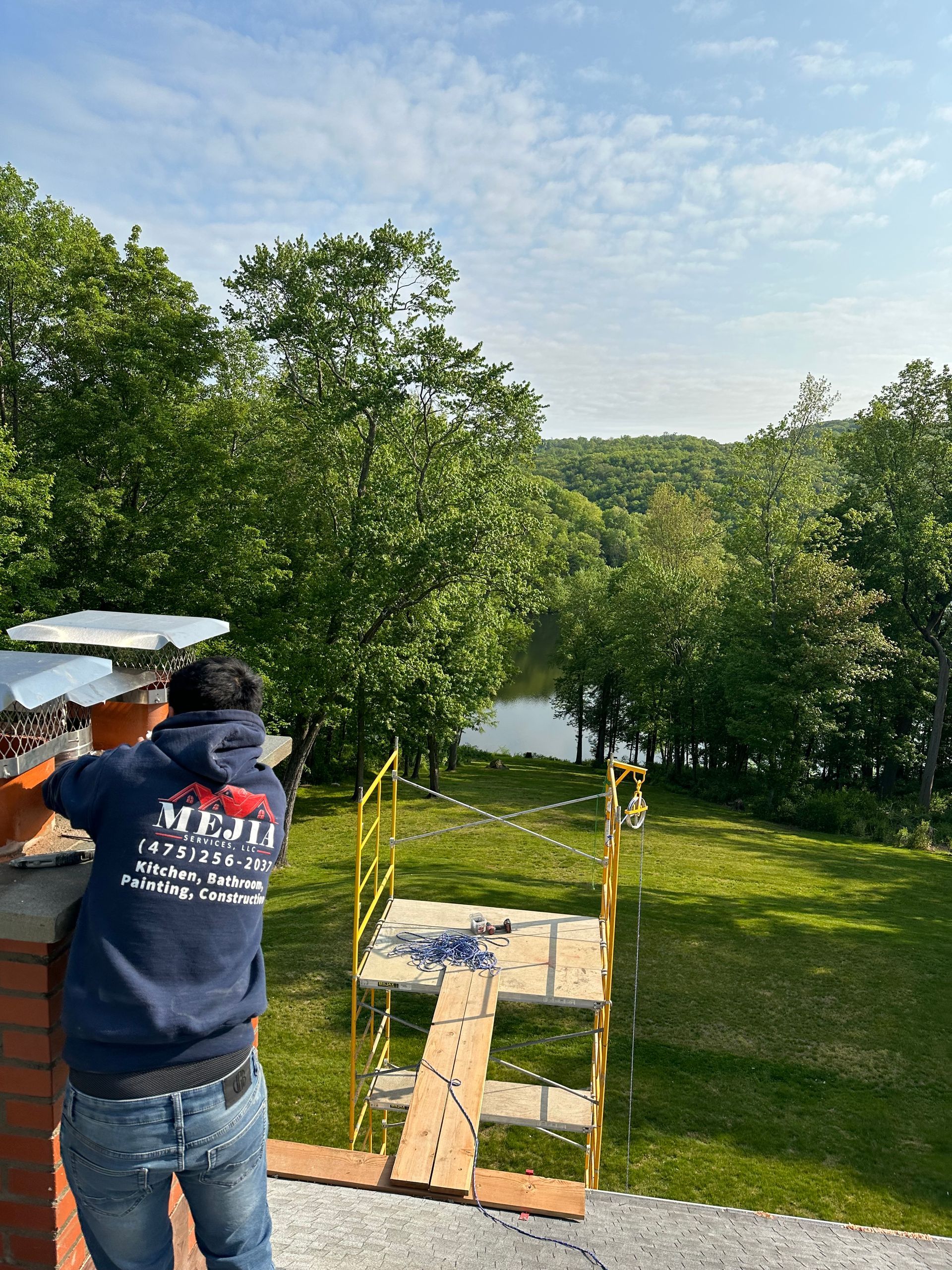 A person in a blue hoodie works on a brick chimney roof overlooking a grassy hill, trees, and a pond.