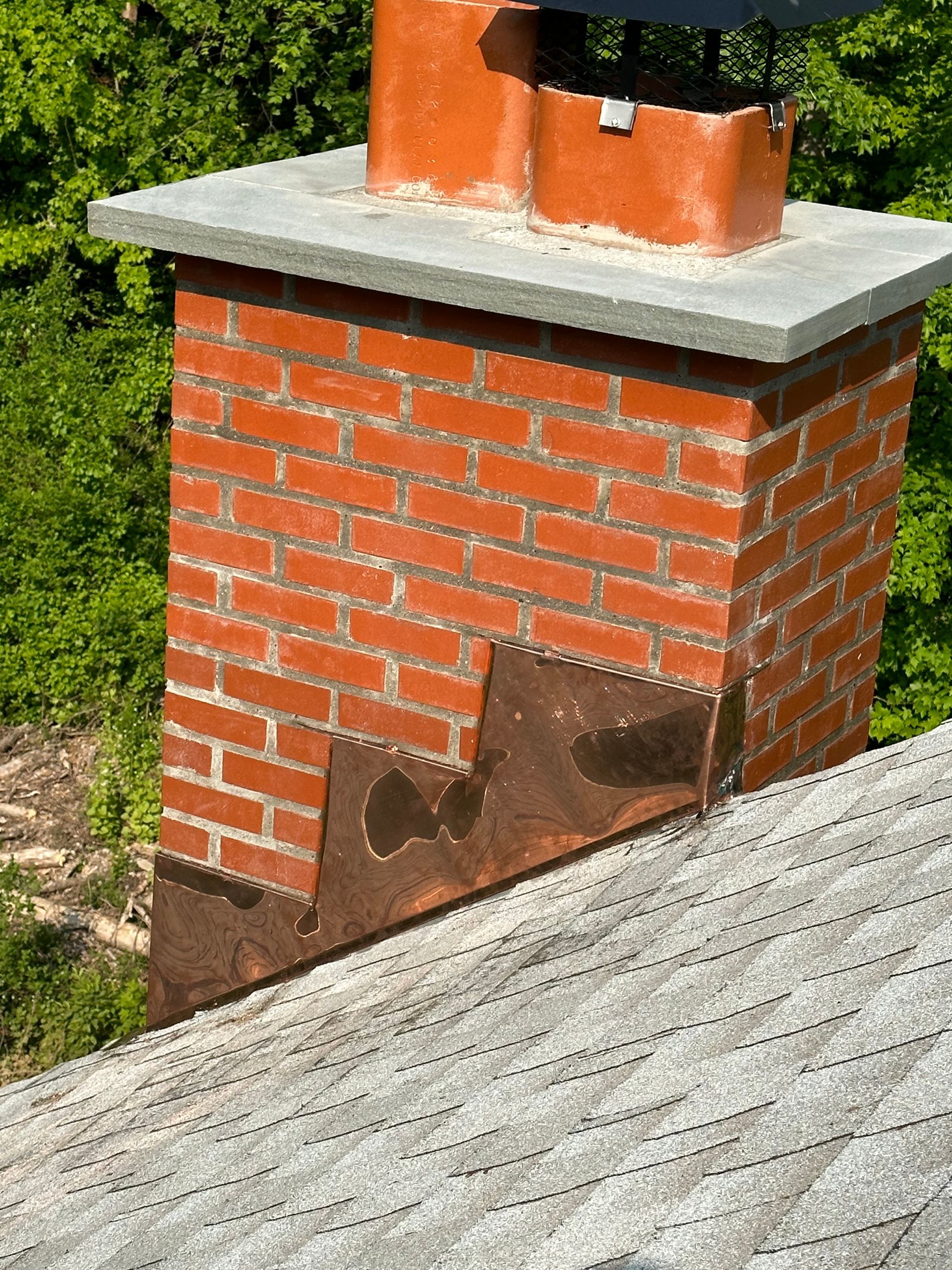 A red brick chimney with a gray stone cap and rusted metal step flashing on a sloped shingled roof.
