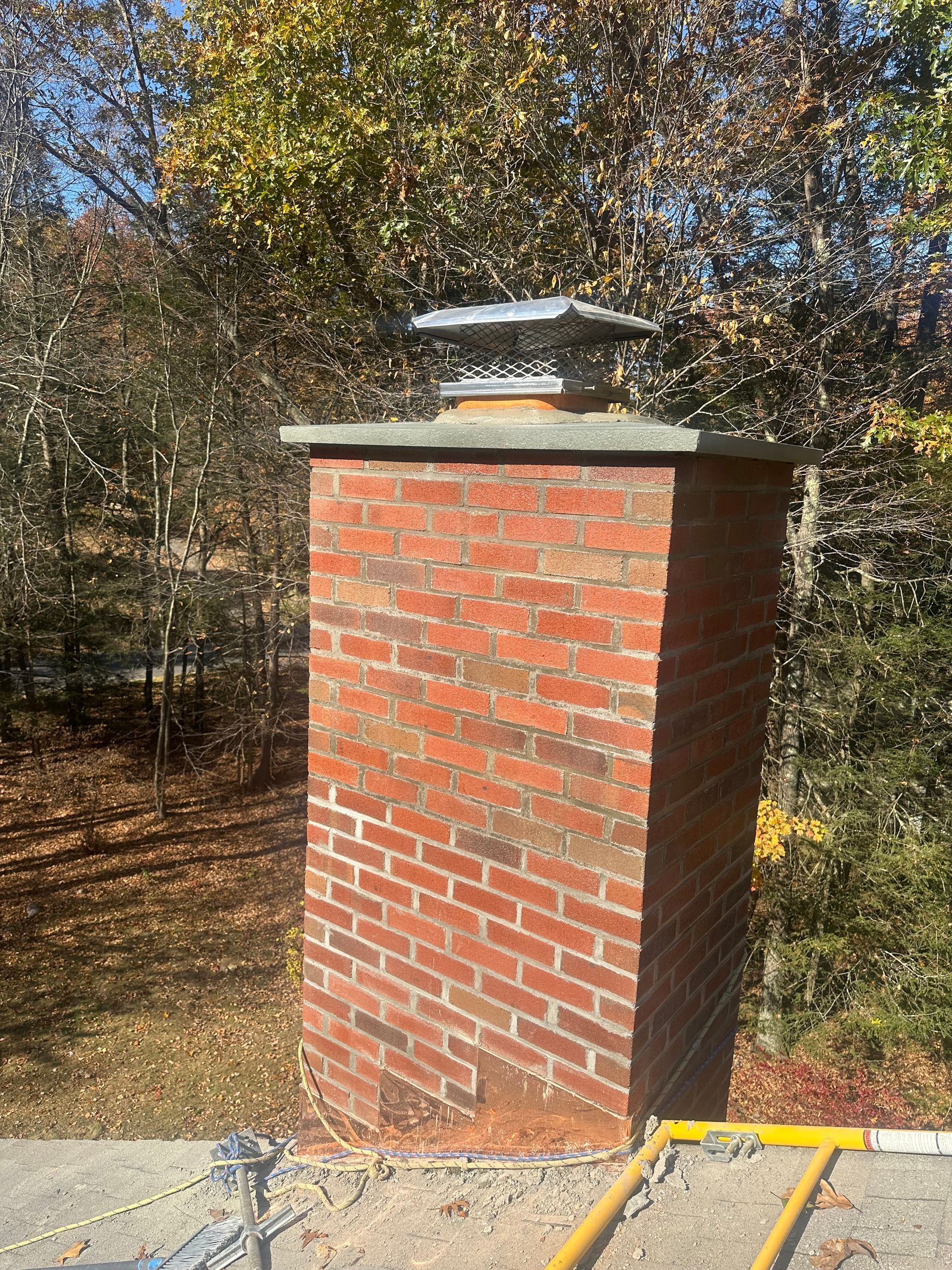 A brick chimney standing on a roof against a background of autumn trees under a bright blue sky.