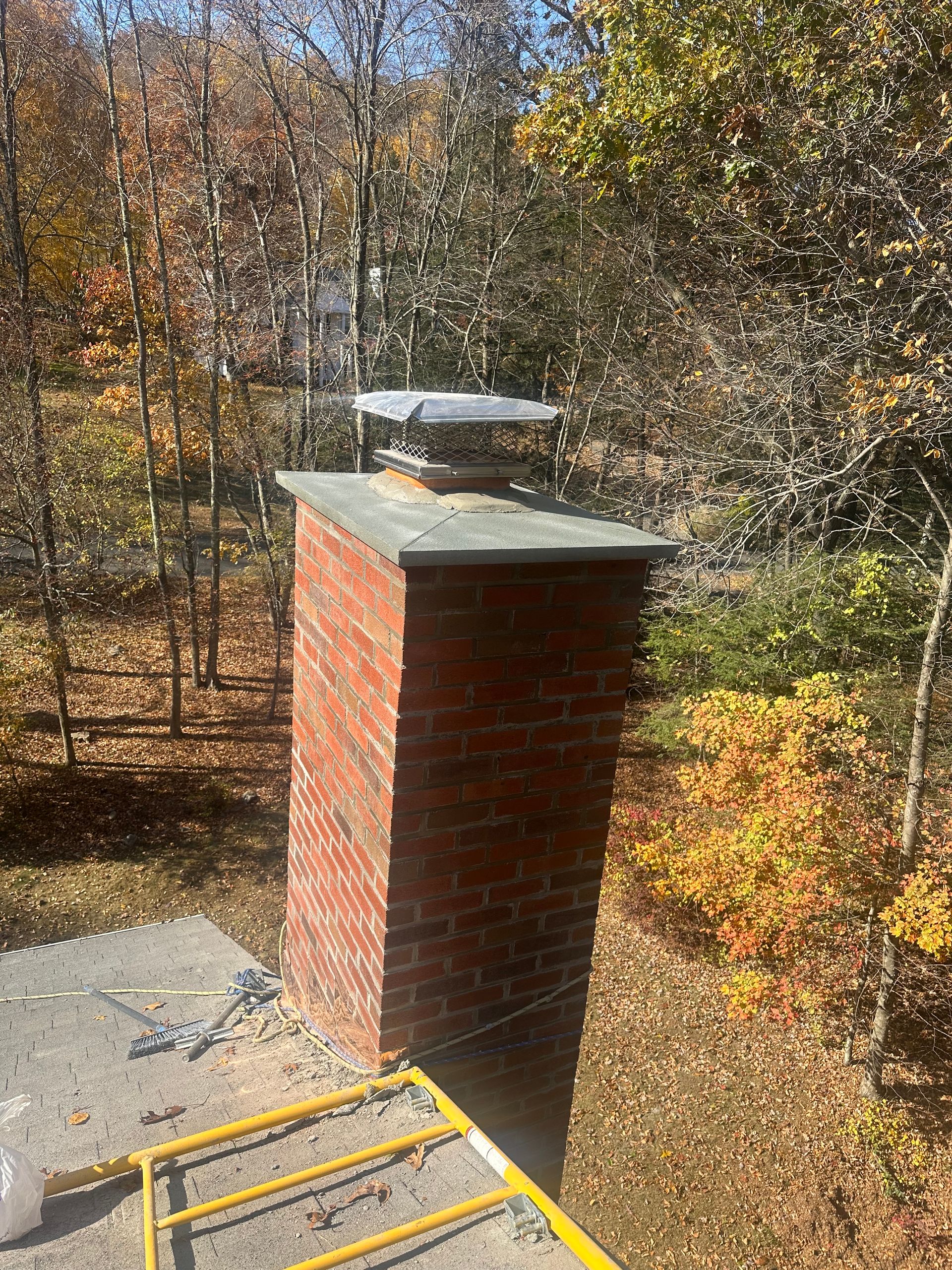 A brick chimney with a metal cap and concrete crown sits on a roof, surrounded by trees with autumn foliage.