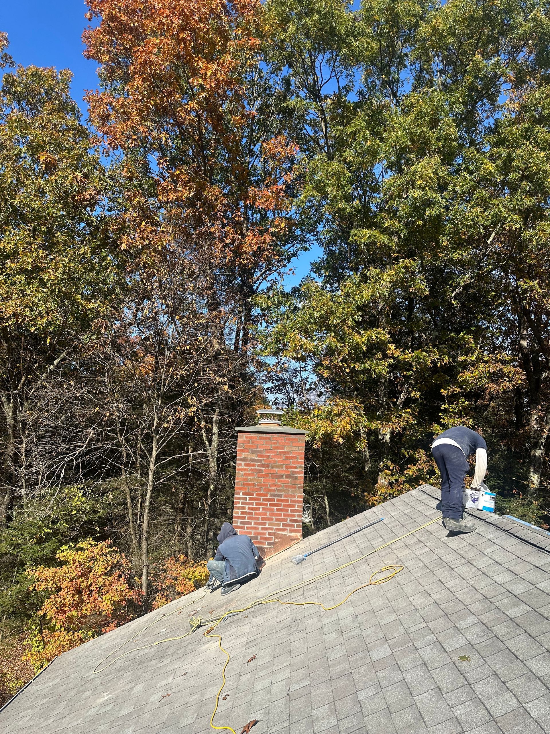 Two people are cleaning leaves off the shingles of a pitched roof next to a brick chimney with autumn trees in the back.