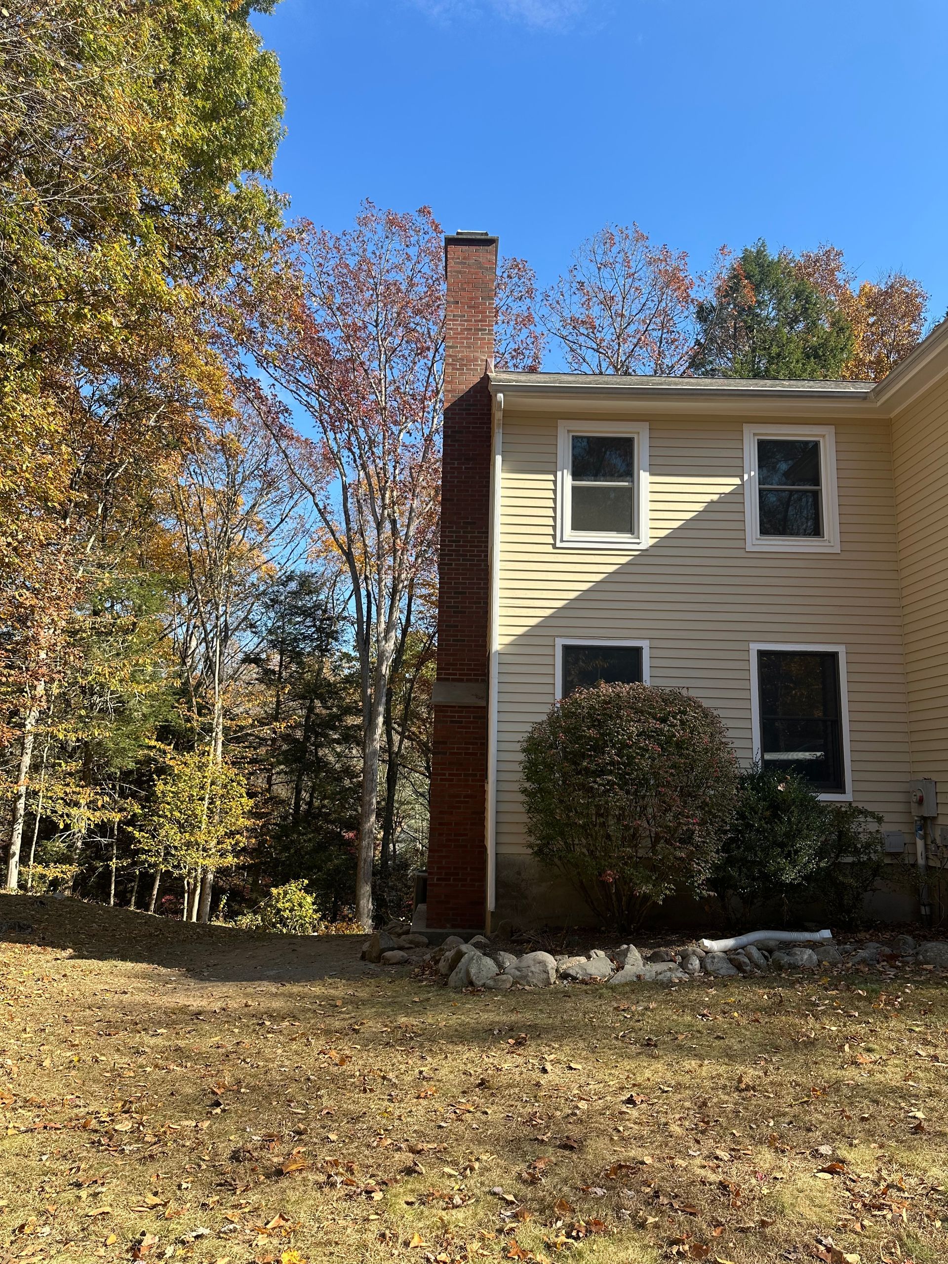 A yellow, two-story house with a brick chimney stands next to trees with autumn leaves under a clear blue sky.