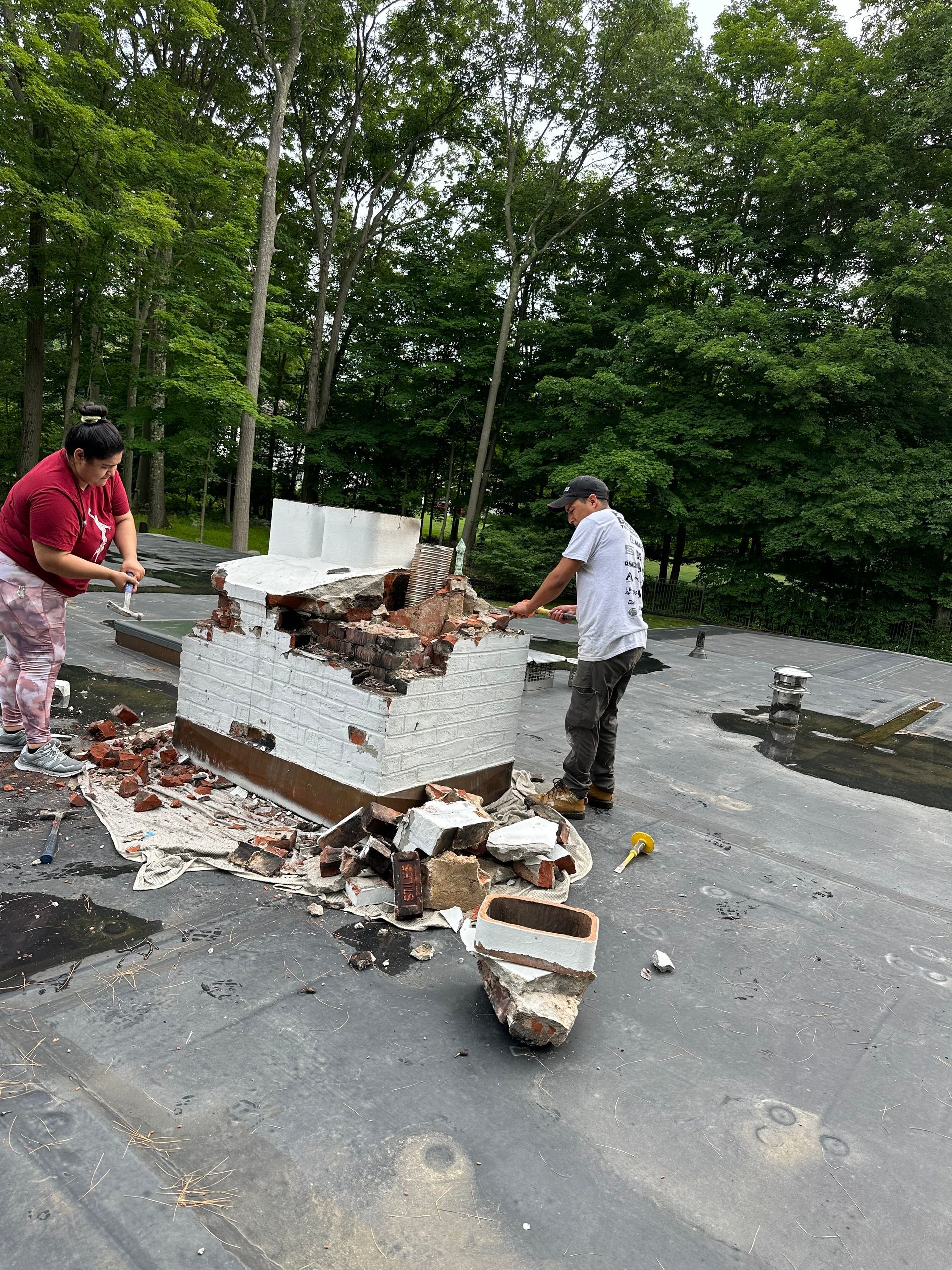 Two people work on demolishing a stone chimney structure on a flat roof surrounded by trees.