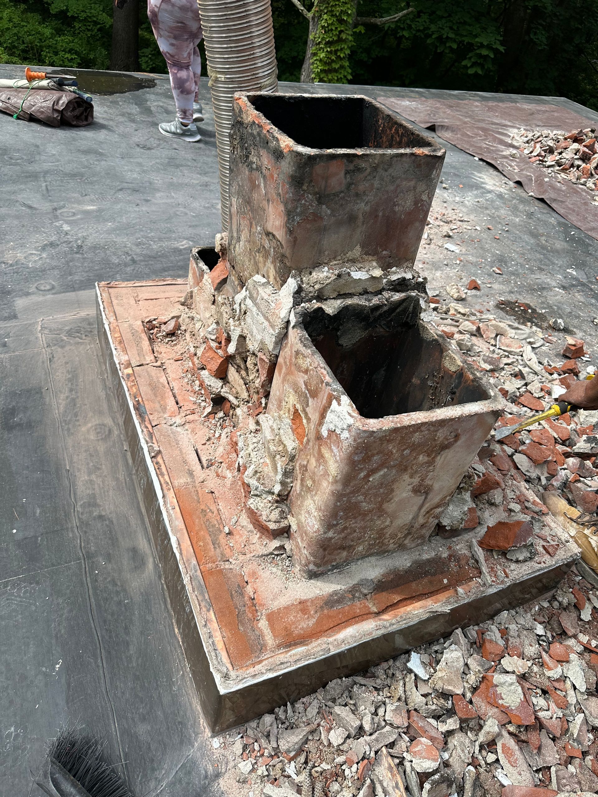 A weathered brick chimney base with two rectangular flue tiles sits on a flat roof surrounded by rubble and construction debris.