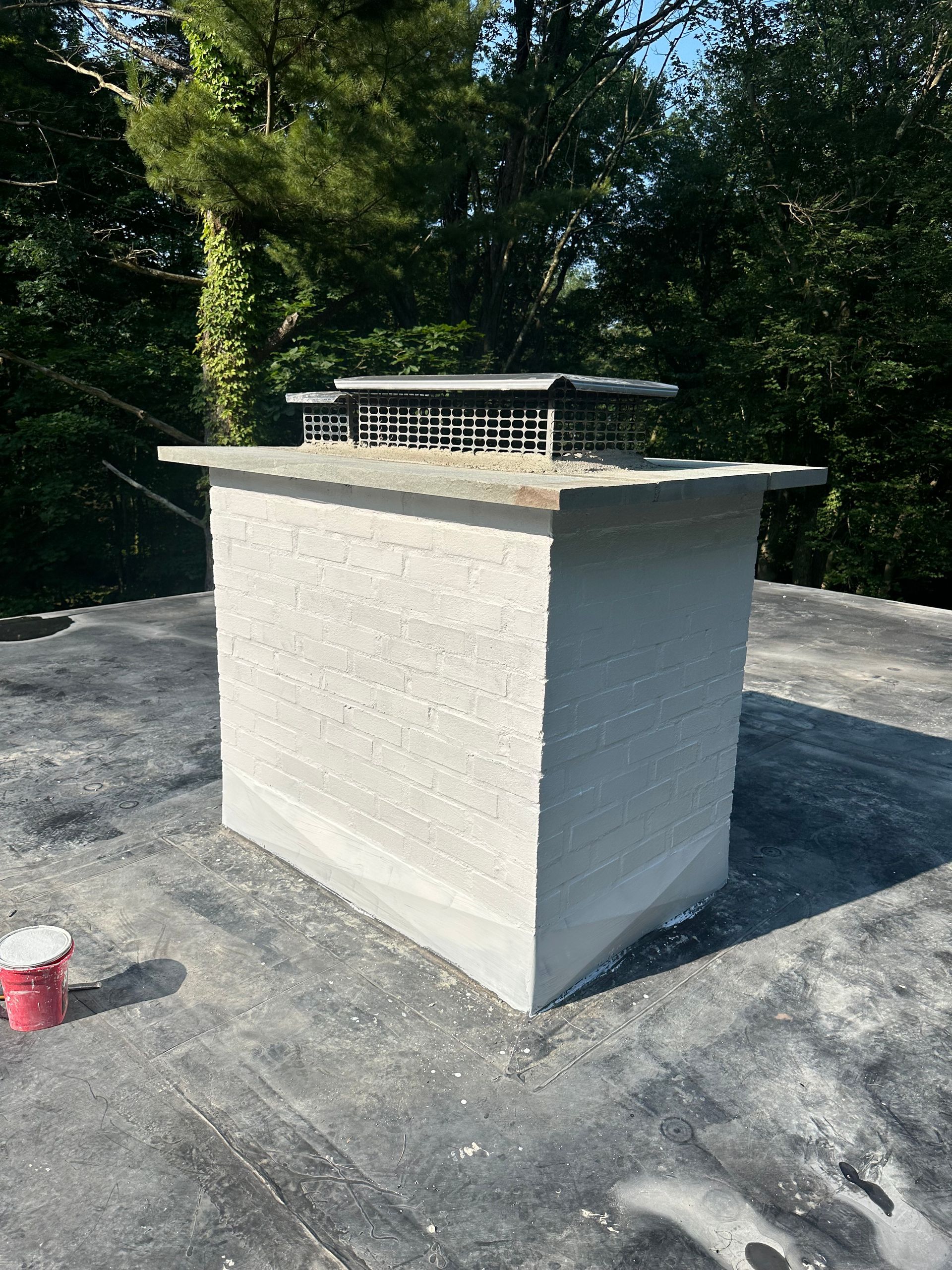 A light-colored, stucco-finished chimney stack with a metal top cage, standing on a flat, dark roof against leafy trees.