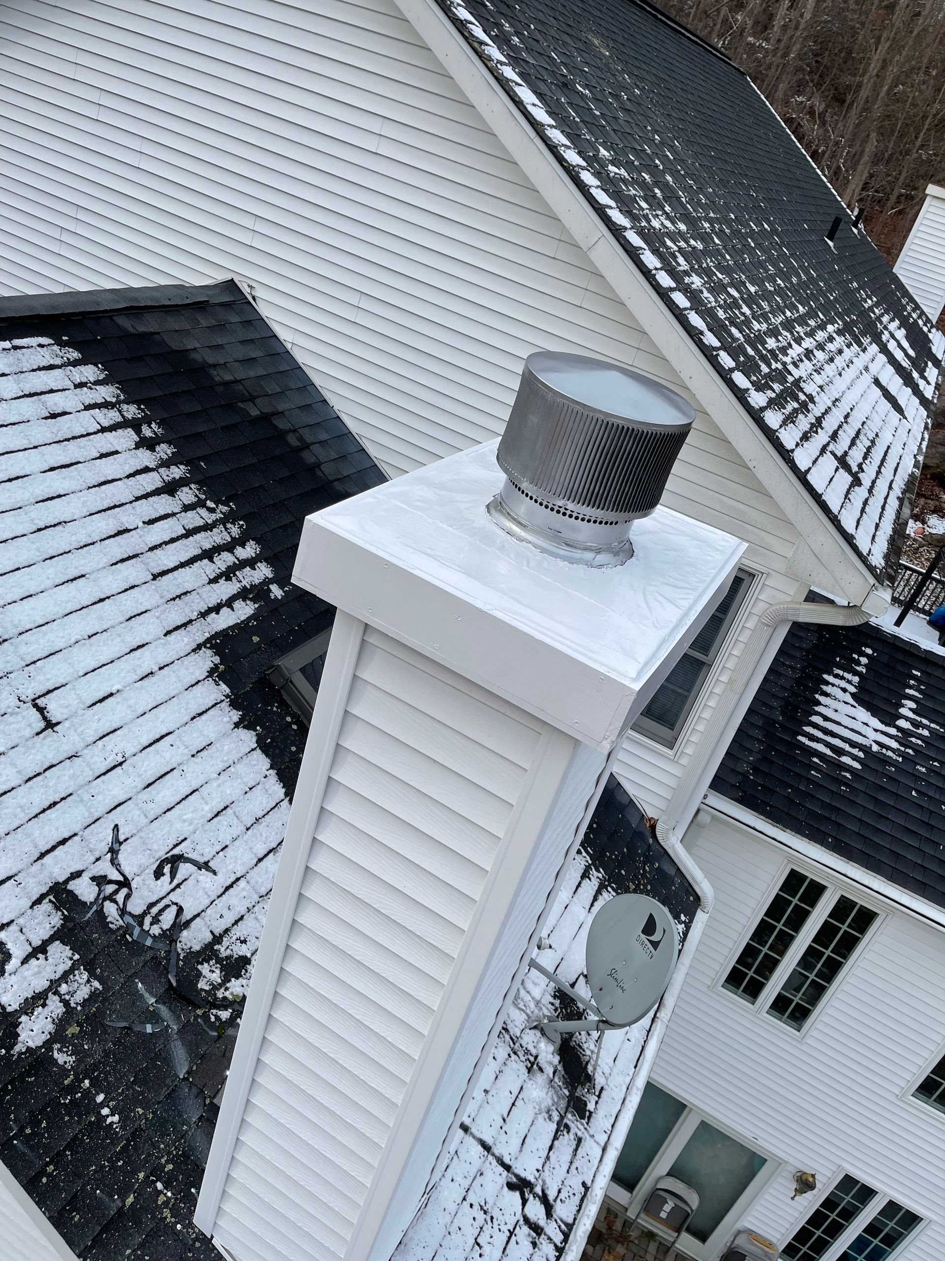 A snow-covered house exterior featuring a tall, white siding chimney with a metallic circular cap on top.