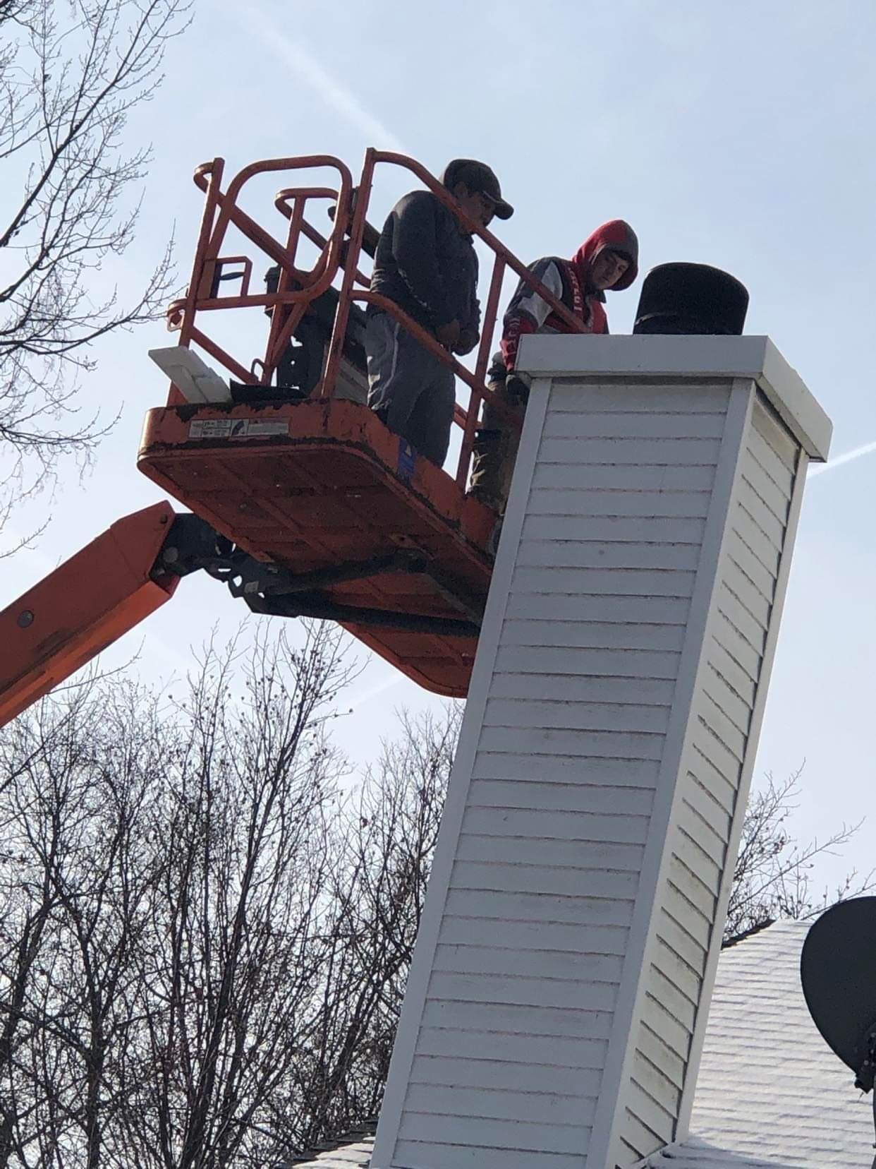 Two workers in an orange aerial lift basket perform repairs on the top of a white, siding-covered chimney on a house roof.