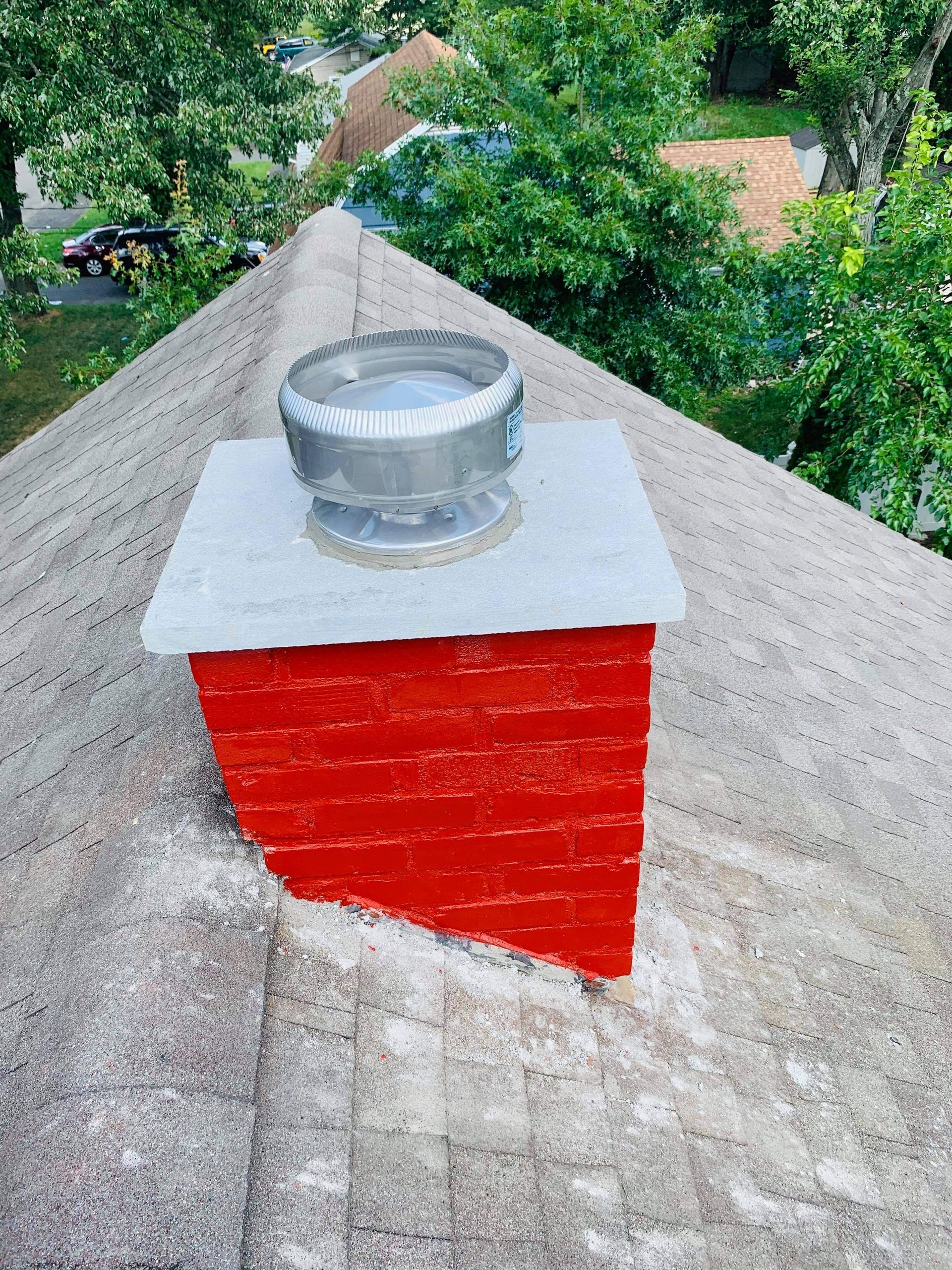 A bright red brick chimney with a metal turbine vent sits on a shingled roof surrounded by green trees.
