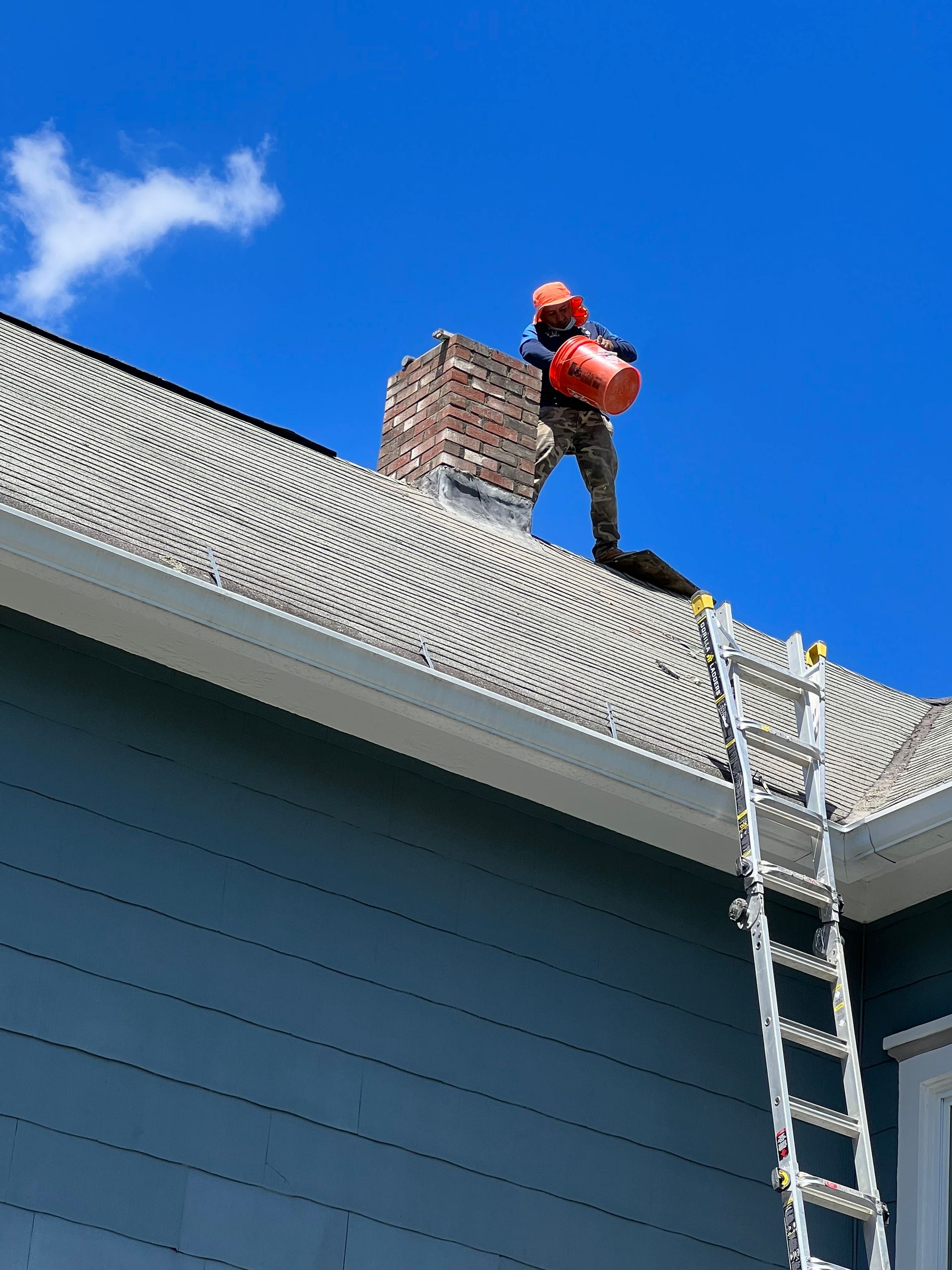 A worker in work gear holds an orange bucket while standing on a shingled roof next to a brick chimney under a blue sky.