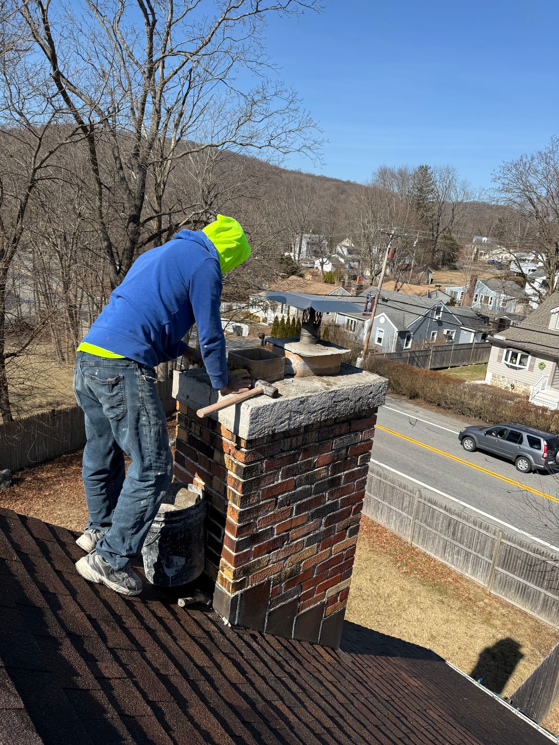 A person in a blue hoodie and neon yellow hat works on a brick chimney on a house roof next to a road.