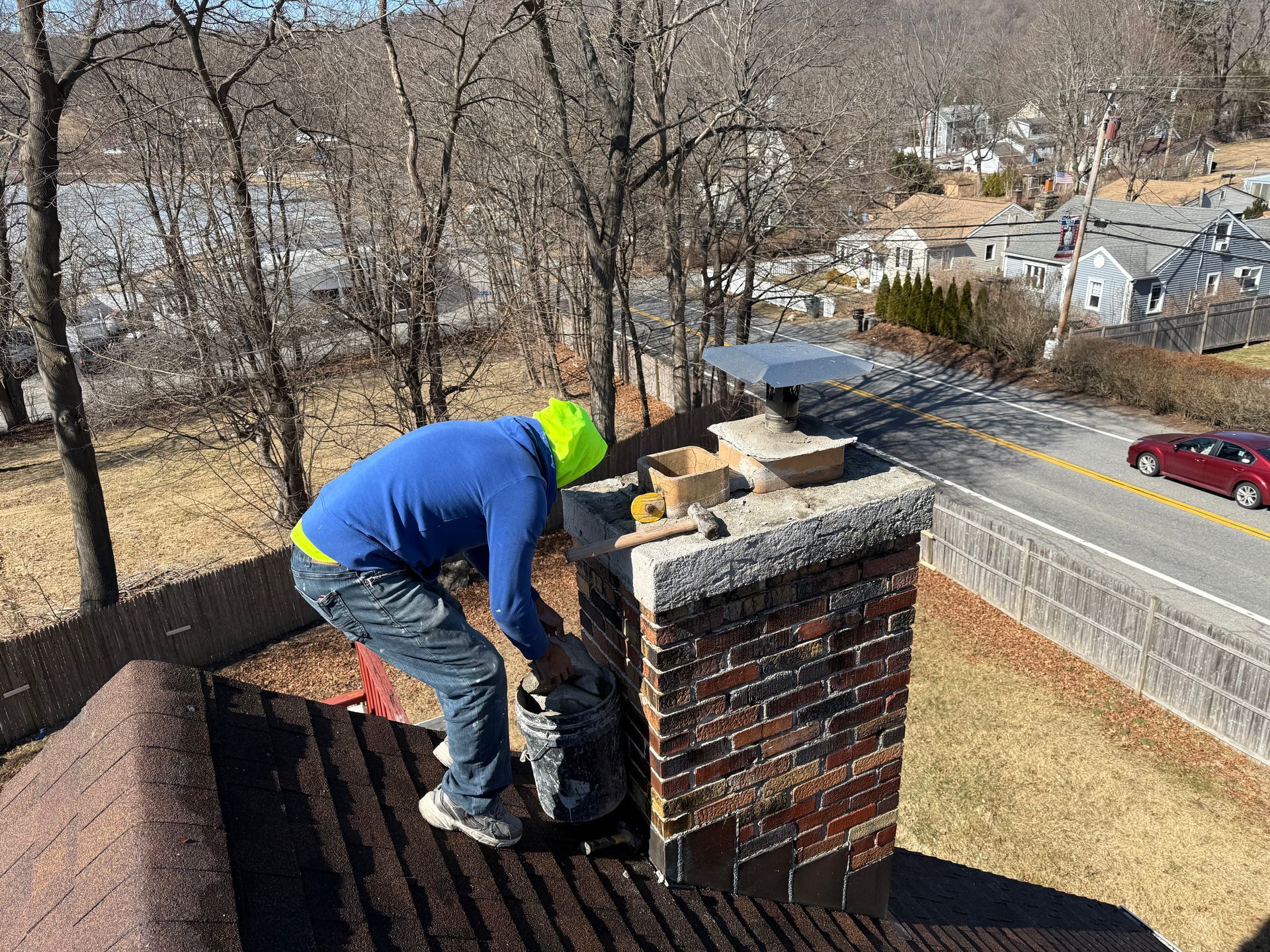 A person in a blue shirt works on a brick chimney on a house roof, with a road and trees visible in the background.