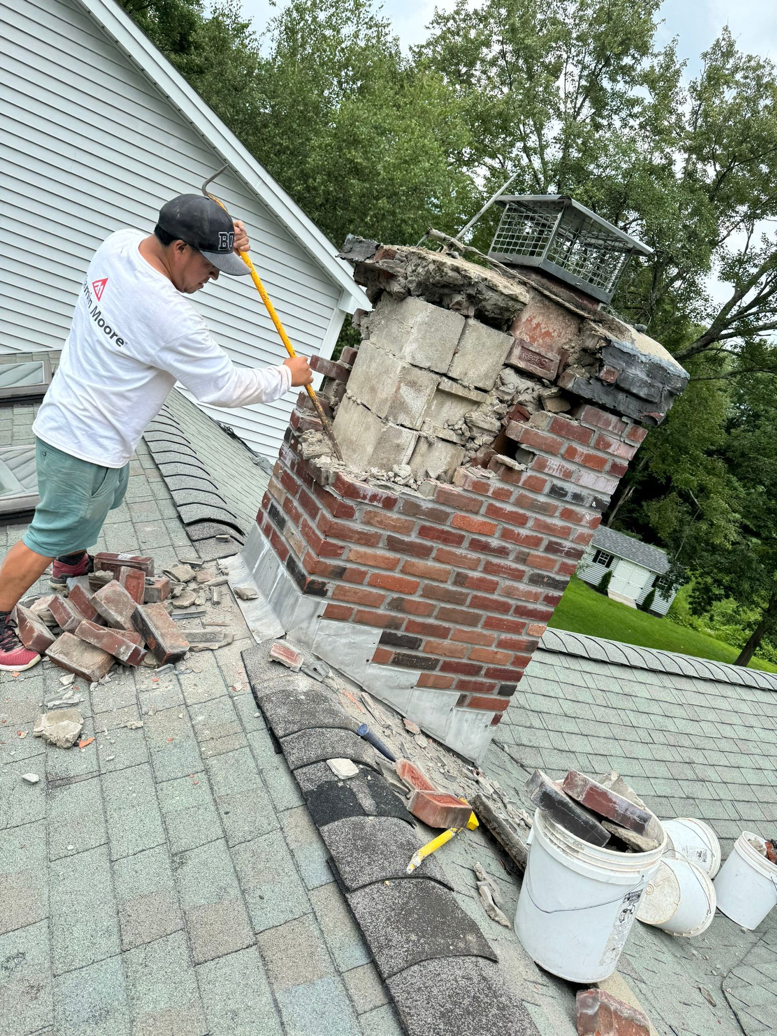 A person wearing a white long-sleeved shirt and shorts uses a hammer to demolish a brick chimney on a shingled roof.
