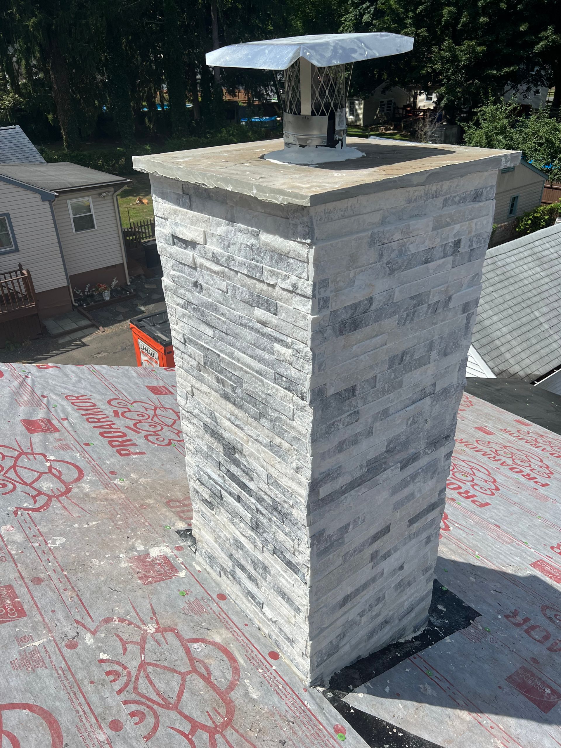A stone-clad chimney with a silver cap sits on a roof covered in red and grey underlayment during construction.
