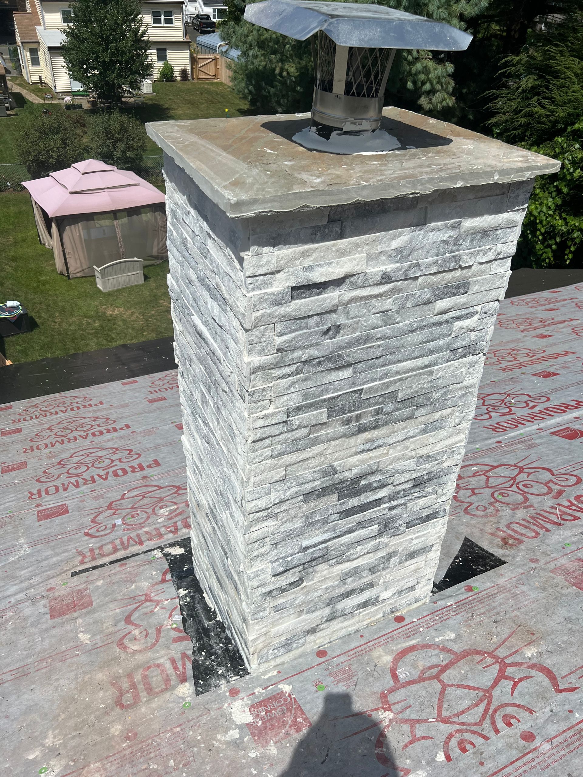 A stone chimney with a metal cap stands on a residential roof covered in underlayment.