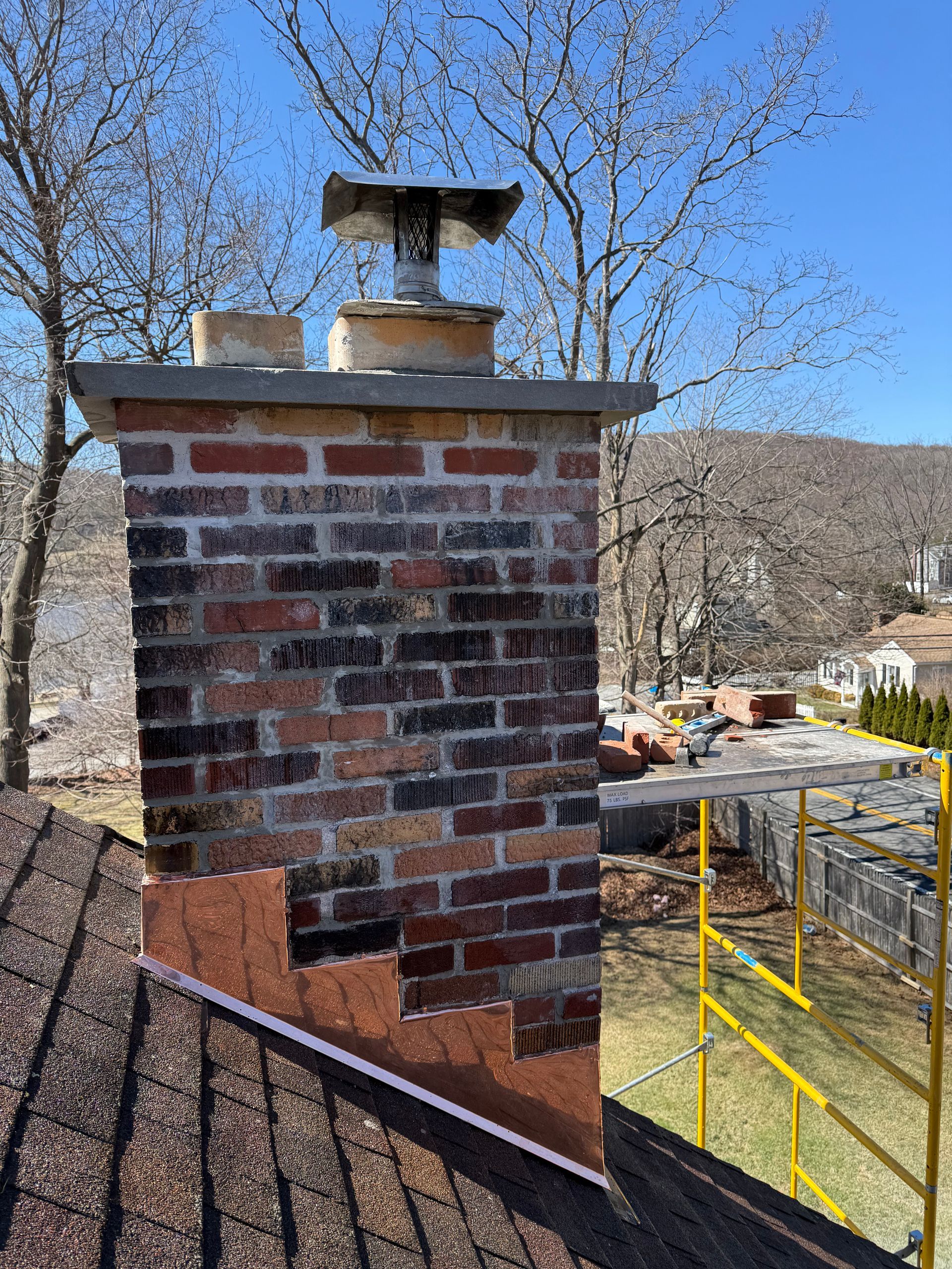 Brick chimney under repair on a shingled roof, featuring exposed underlayment and scaffolding nearby against a blue sky.