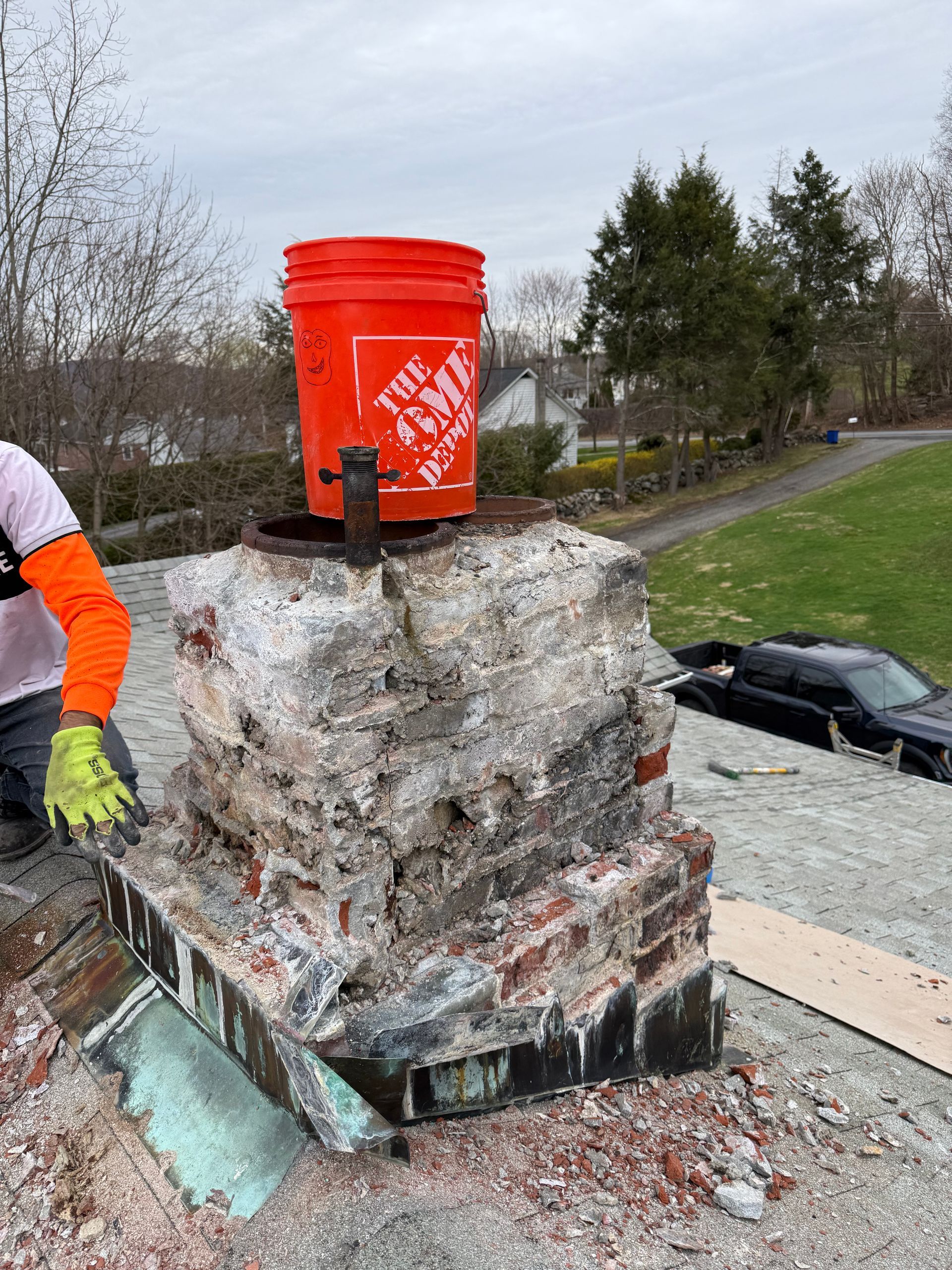 A worker in bright orange sleeves and gloves repairs a crumbling brick chimney on a roof with an orange bucket on top.