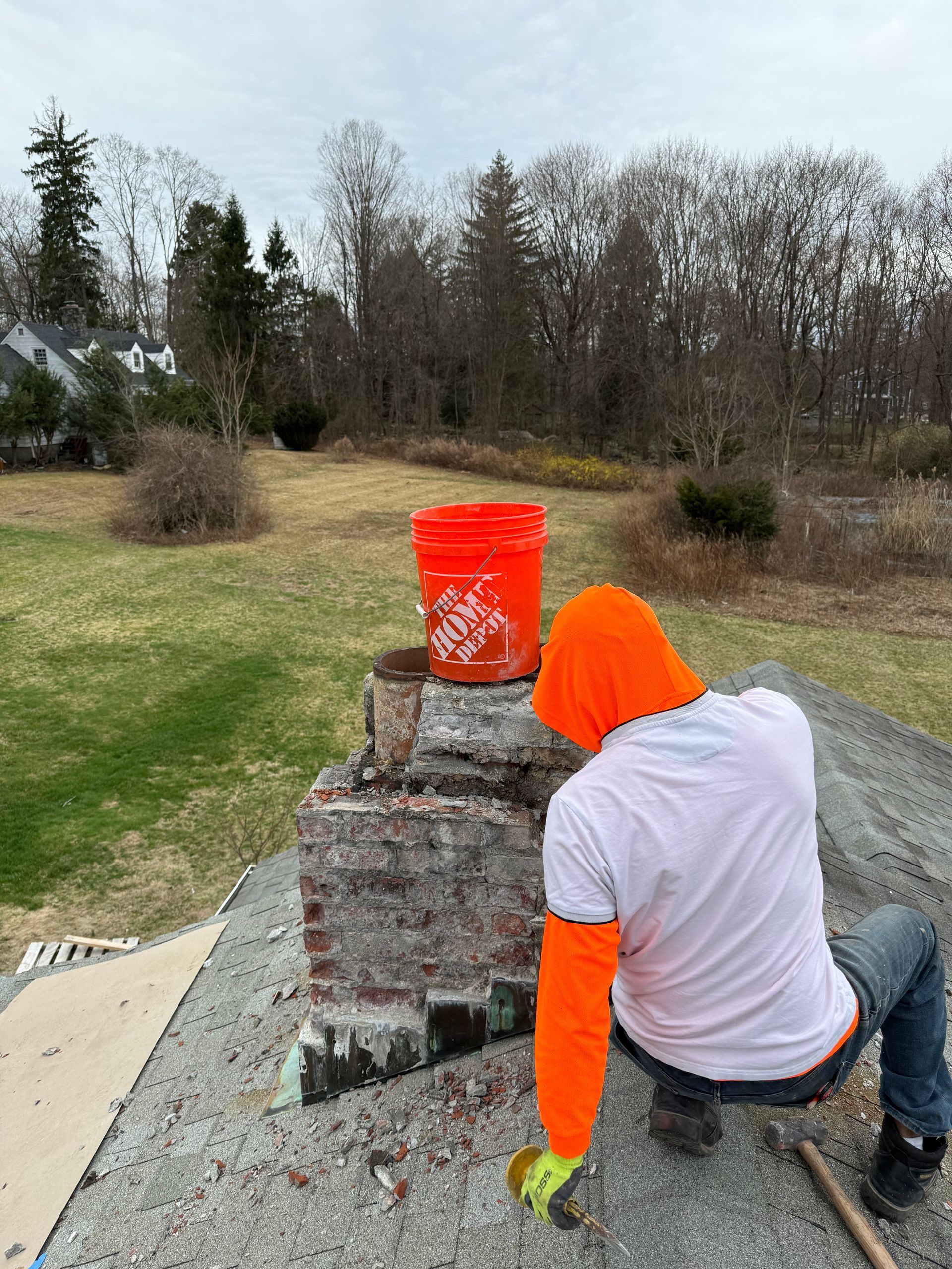A person wearing an orange hoodie and safety sleeves kneels on a roof, working on a brick chimney with an orange bucket.