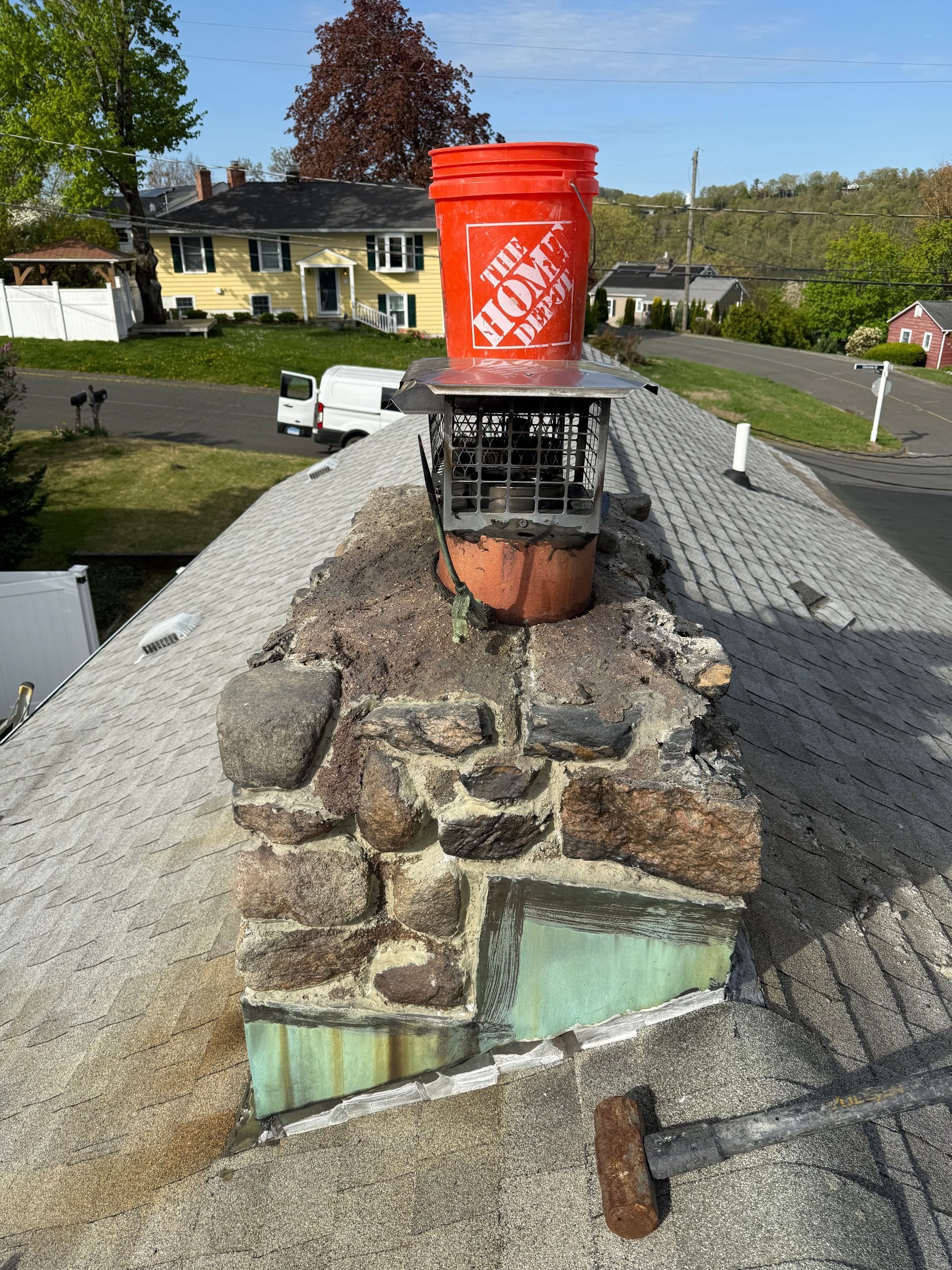 An orange Home Depot bucket sits upside down on a metal chimney cap atop a stone chimney on a shingled roof.