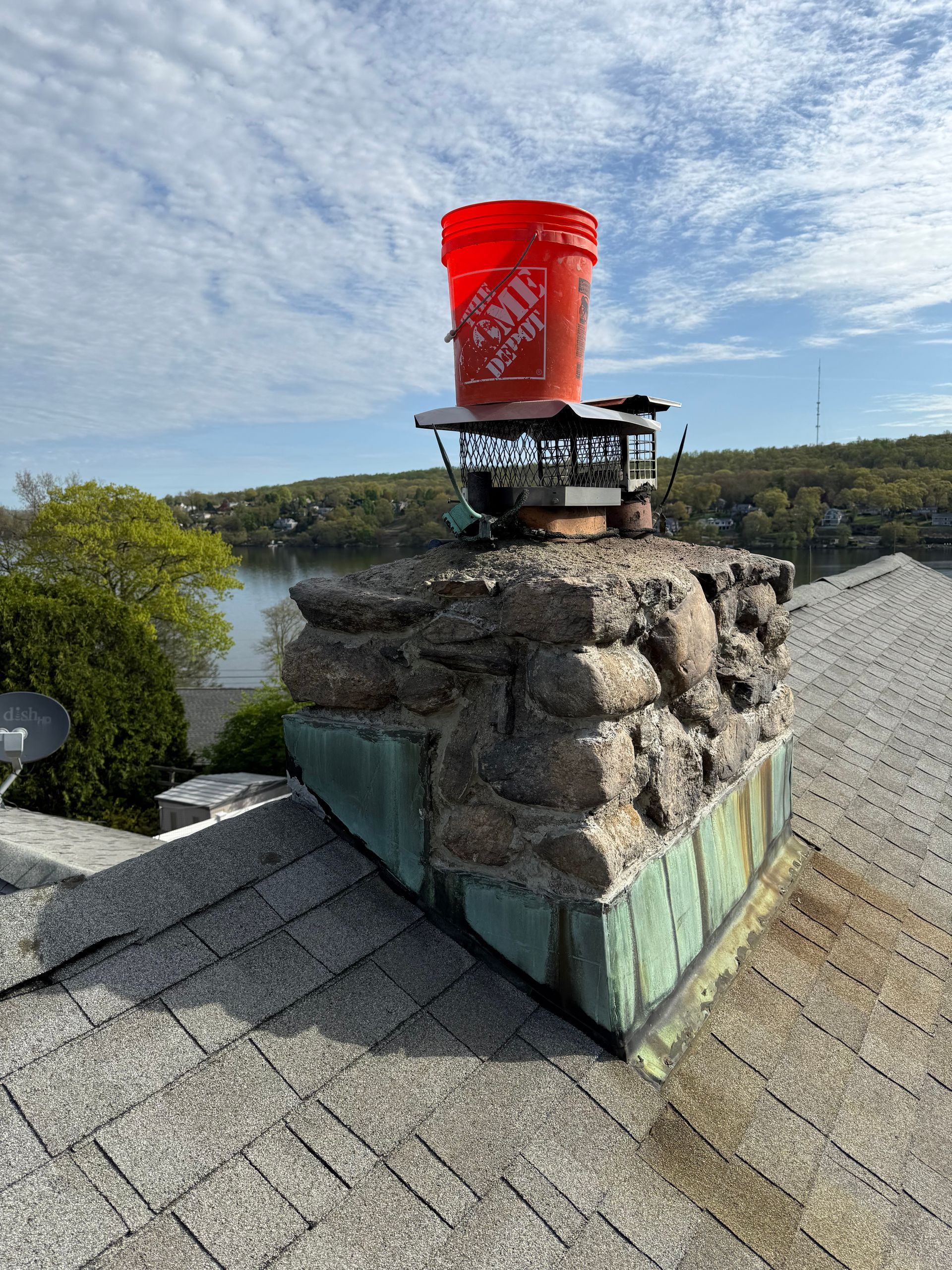 A red Home Depot bucket sits upside down on top of a stone chimney cap on a shingled roof with a lake in the background.
