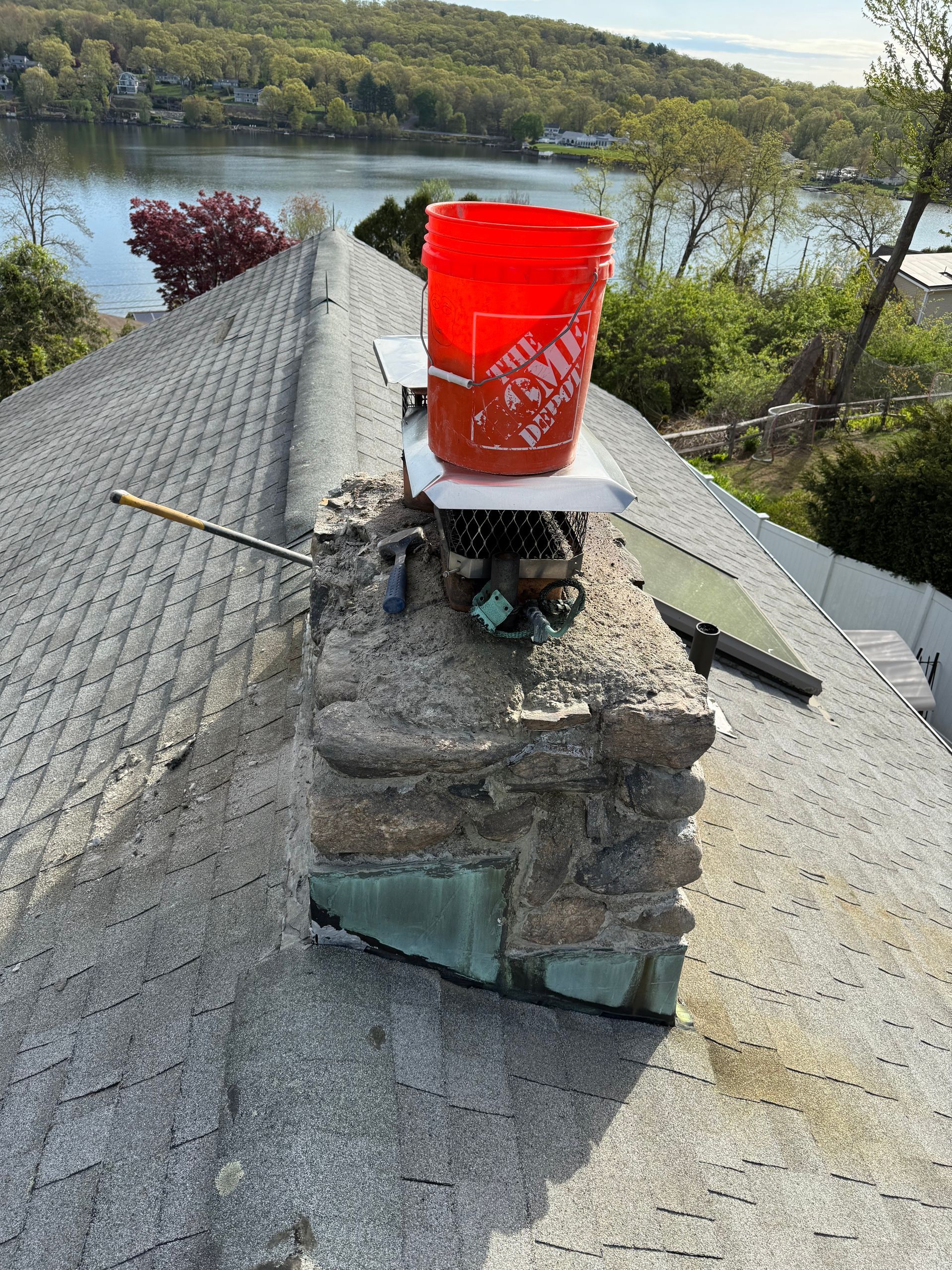 A bright orange Home Depot bucket sits on top of a weathered stone chimney on a sloped roof overlooking a lake.