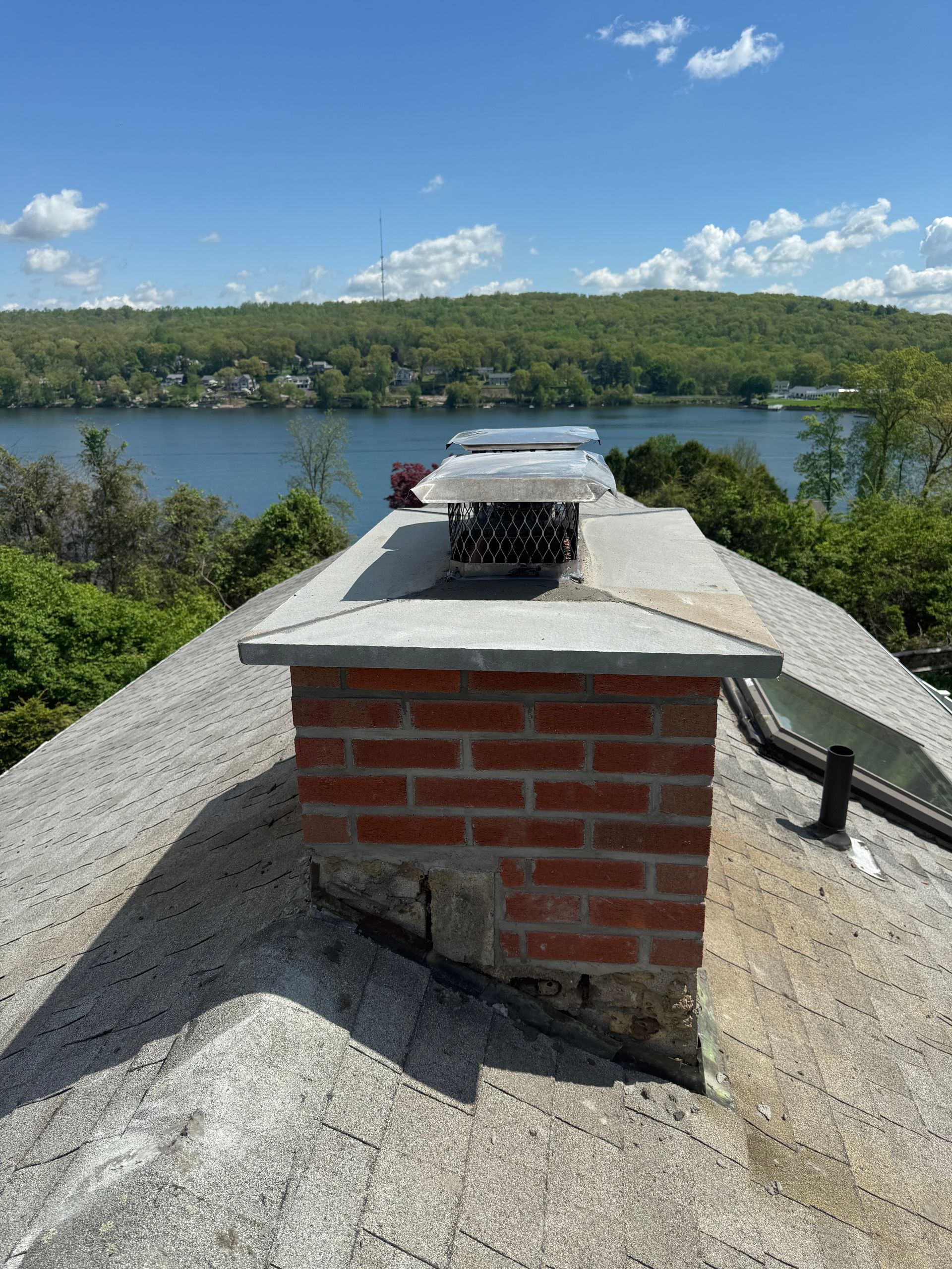 A brick chimney with a metal cap stands on a shingled roof overlooking a lake and forested hills under a blue sky.