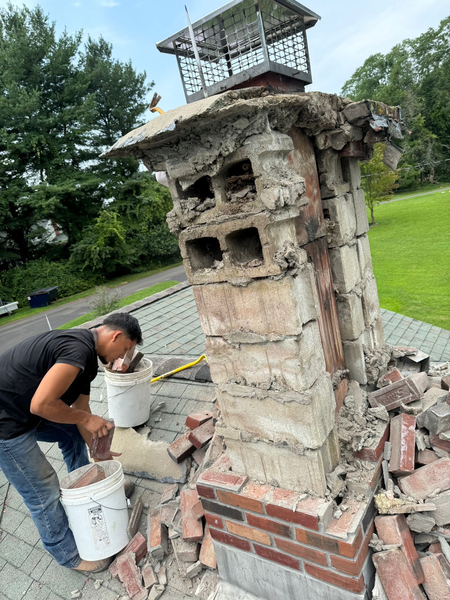 A worker in a black shirt repairs a damaged brick chimney on a shingled roof, putting debris into a white bucket.