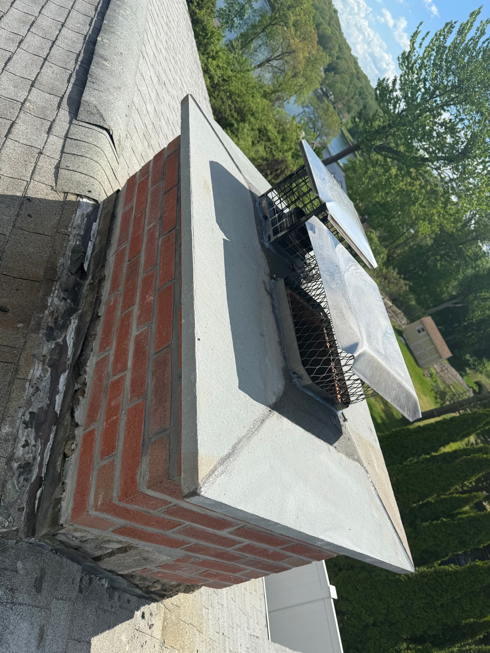 A brick chimney on a shingled roof features a light-colored concrete cap and two metal flue covers.