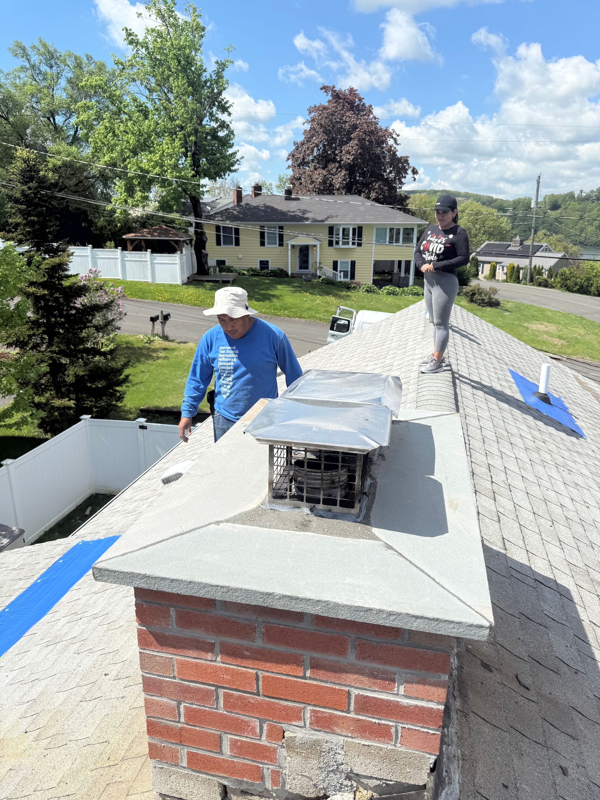 Two people work on a residential roof featuring a brick chimney, with a suburban neighborhood in the background.