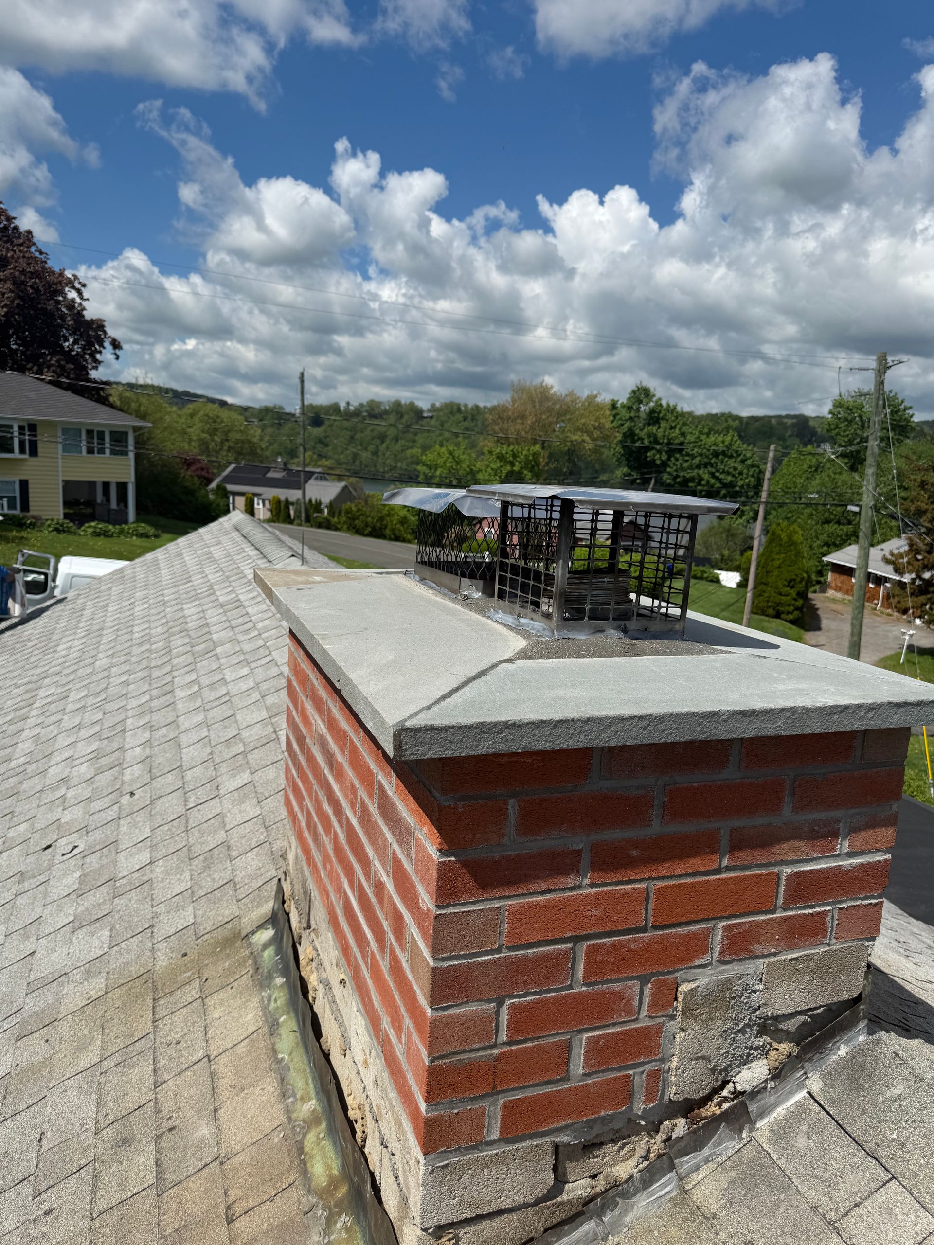 A brick chimney on a shingled residential roof with a metal cap, overlooking a green landscape under a cloudy blue sky.