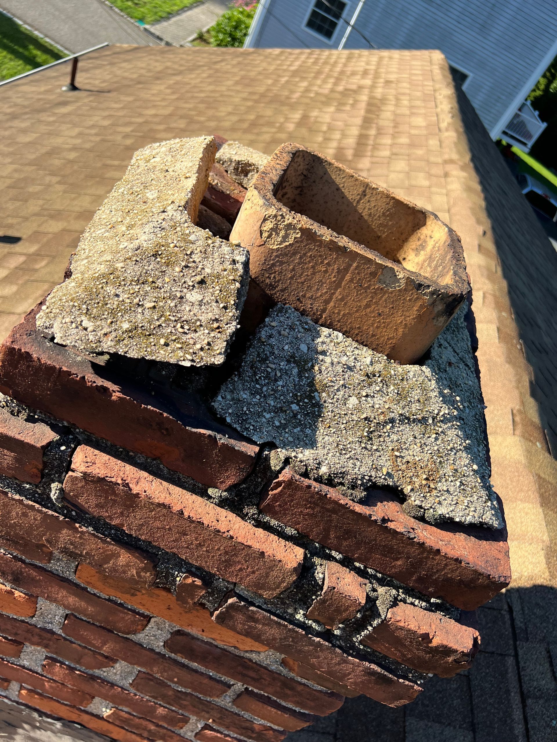 A brick chimney top with a broken concrete crown and a loose terracotta flue liner on a brown shingled roof.