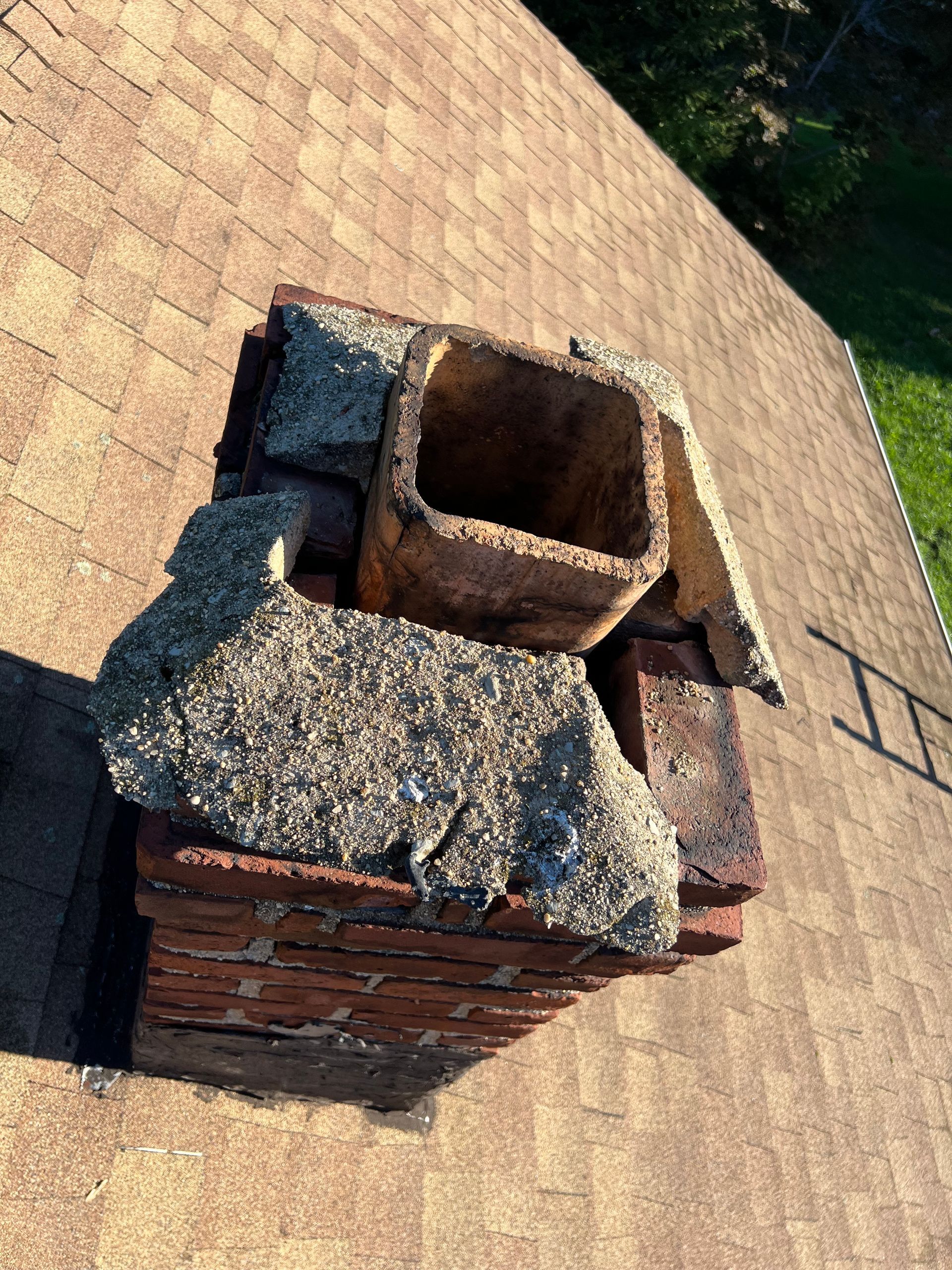 A crumbling concrete chimney crown with an exposed clay flue liner, situated on a shingled roof.