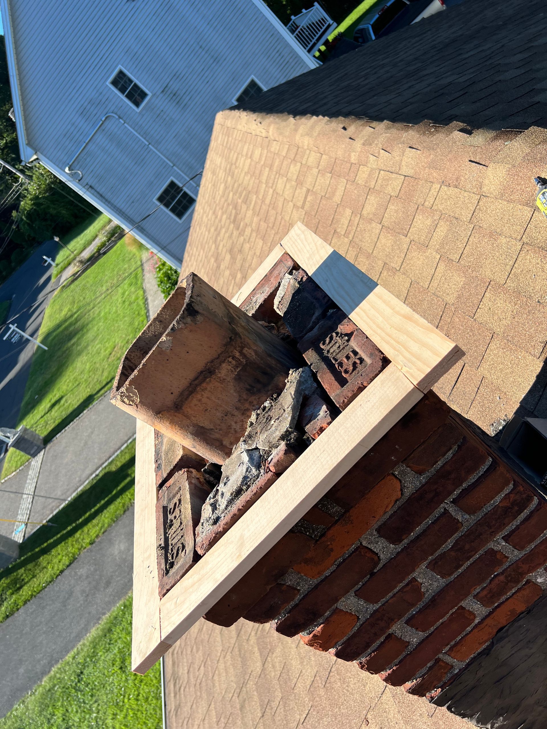 A damaged brick chimney on a shingled roof with a temporary wooden frame placed around its top opening.