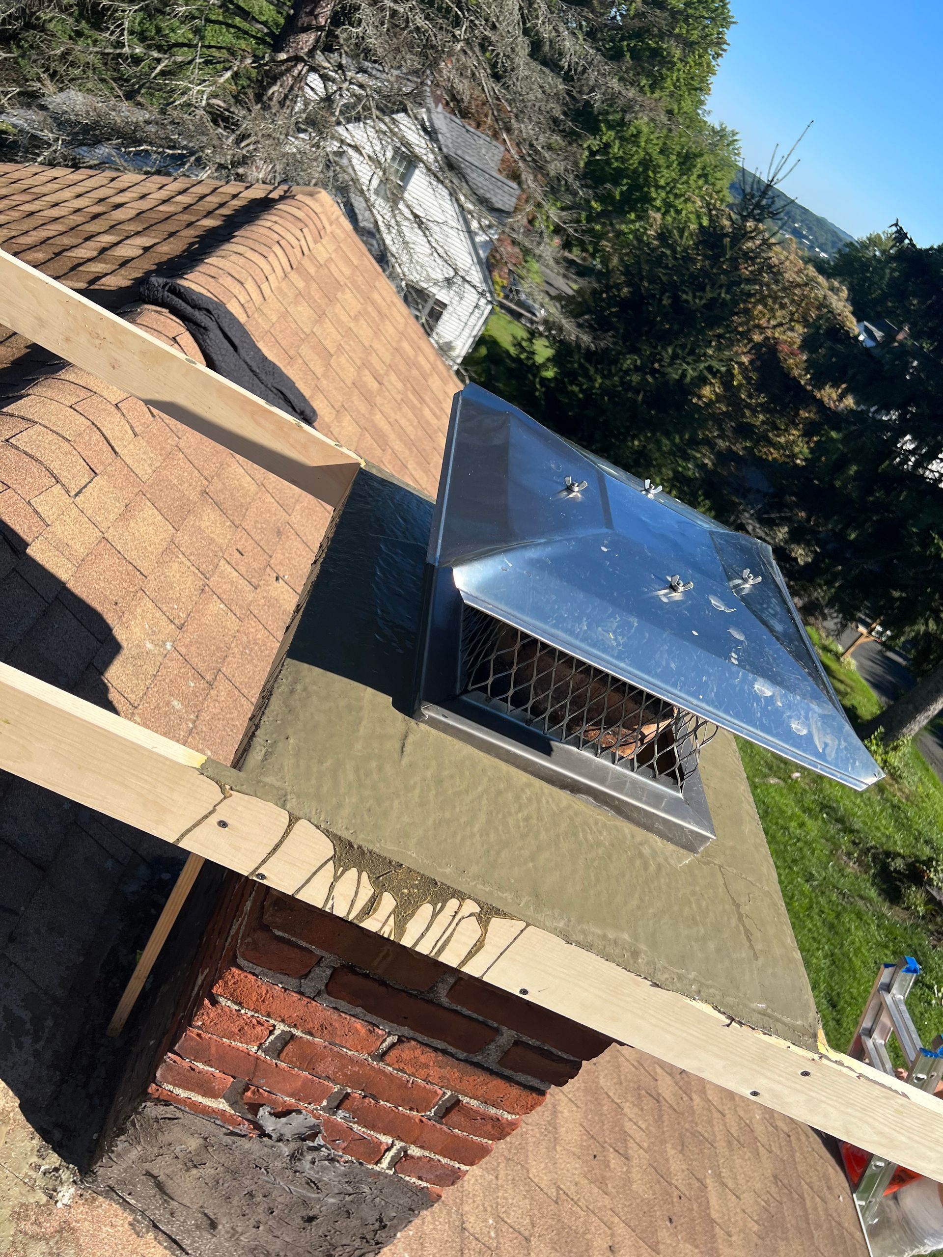 A high-angle view of a brick chimney on a roof under construction, featuring a new metal chimney cap and wooden framing.