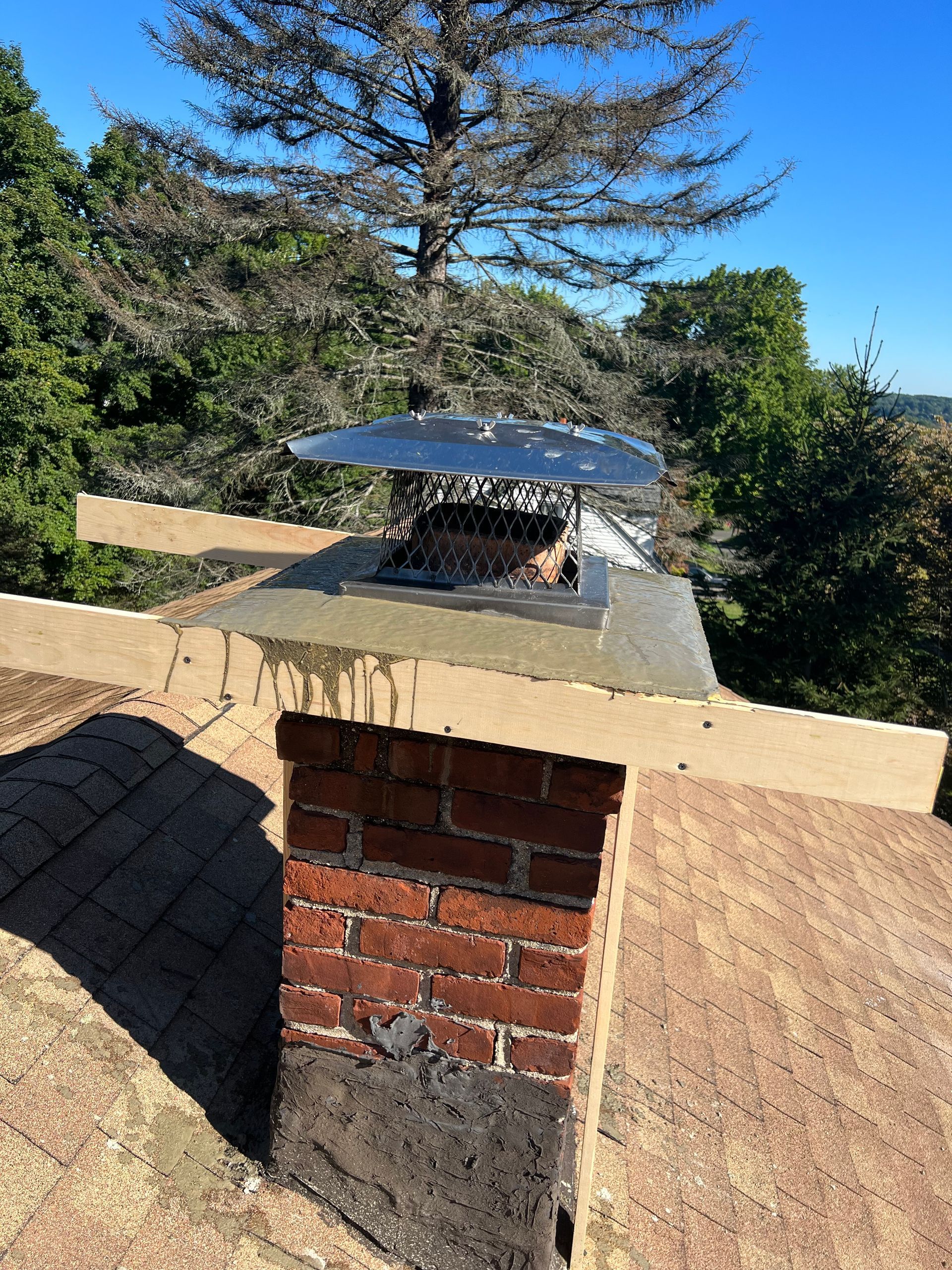 A red brick chimney on a shingled roof with a new concrete crown and a stainless steel chimney cap.