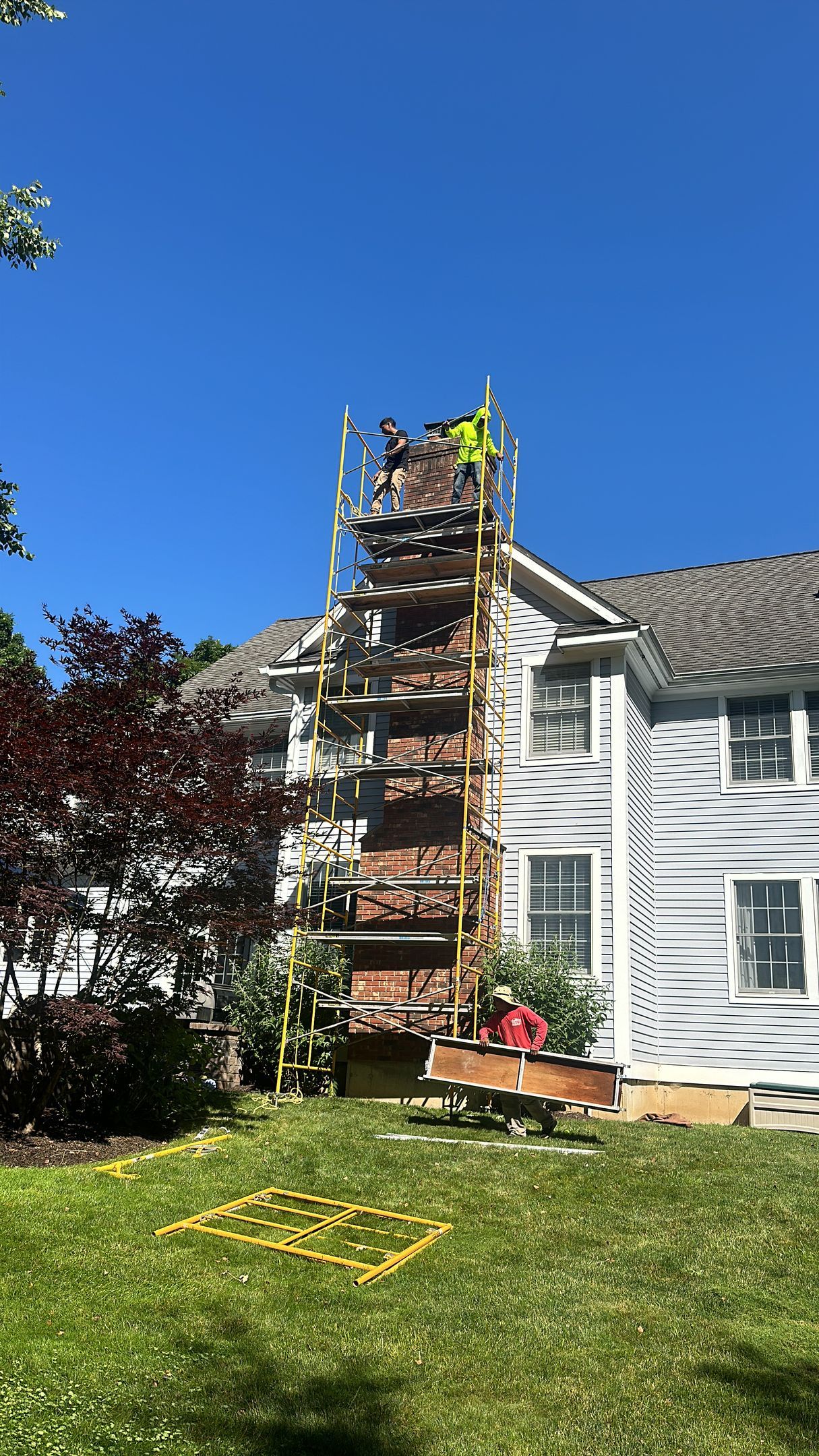 Workers on high scaffolding performing chimney repairs on the exterior of a white house on a sunny day.