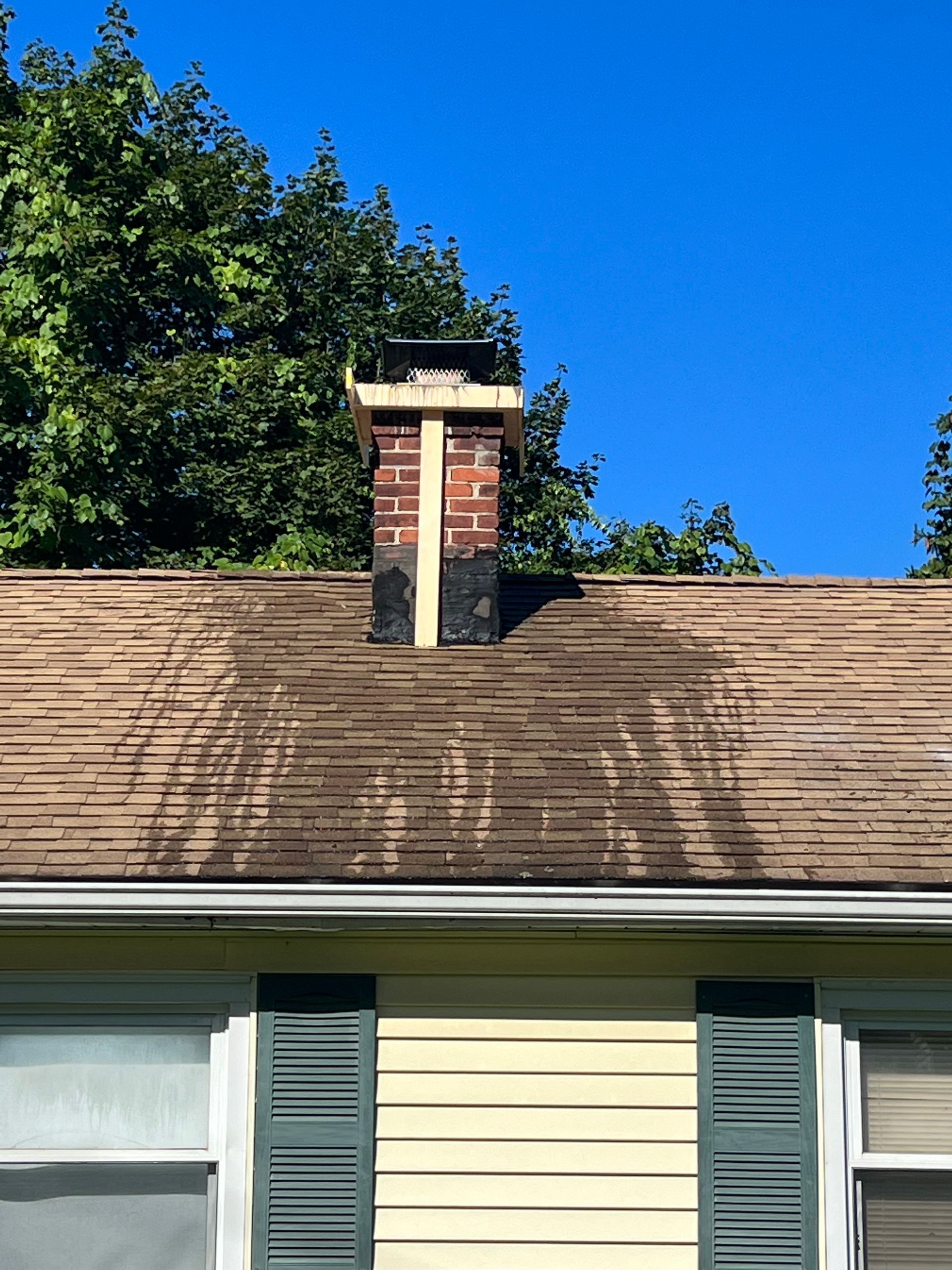 A brick chimney with a wooden support frame on a brown shingled roof under a clear blue sky.